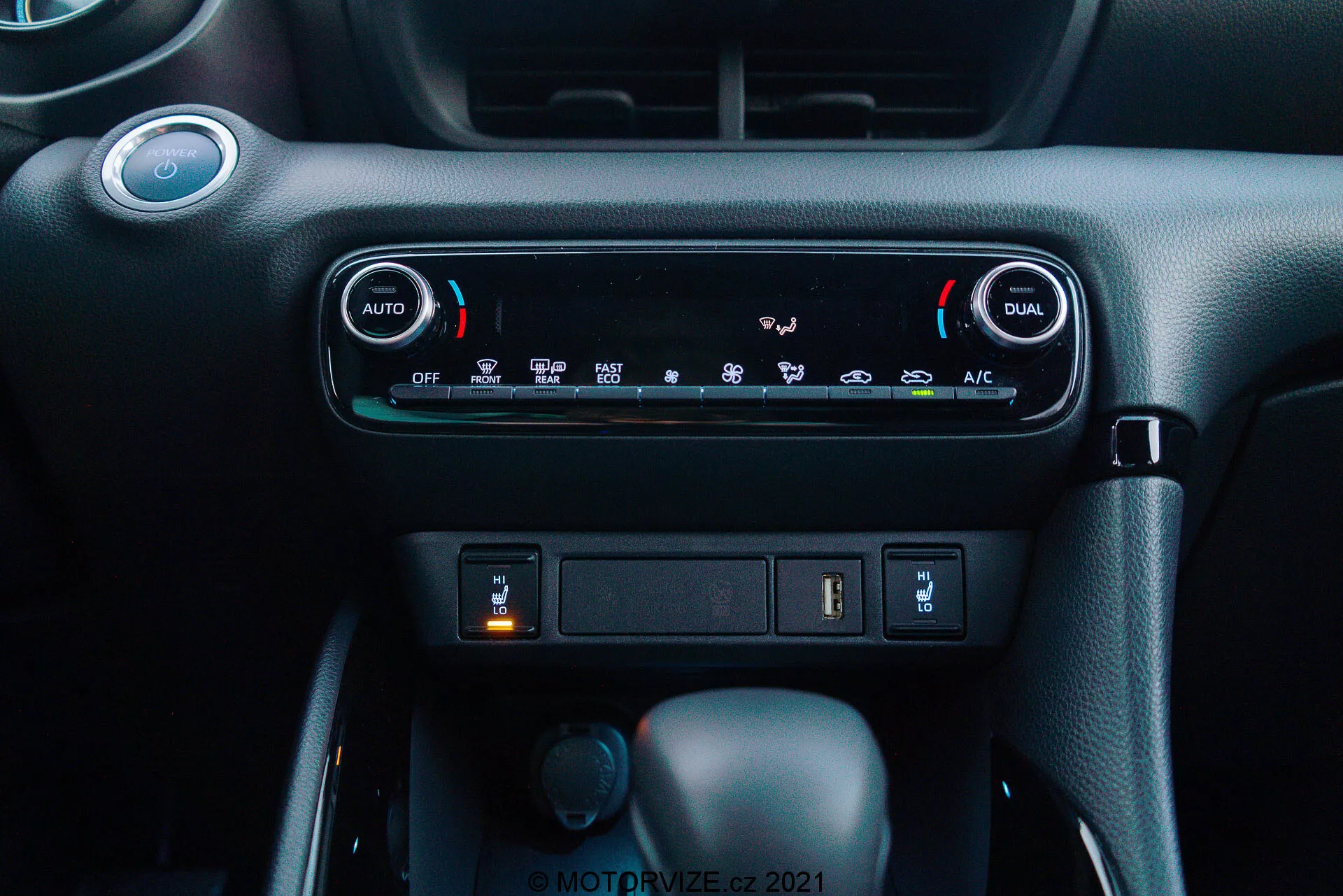 Vue rapprochée du tableau de bord de la Toyota Yaris 2020 : L'image met en avant le système de climatisation à double zone automatique, avec des boutons 'AUTO' et 'DUAL' pour des réglages de température indépendants pour le conducteur et le passager. Sous les commandes de climatisation, un compartiment de rangement avec un port USB et une éventuelle prise de courant de 12V est visible. En dessous, trois boutons sont visibles : le bouton central est le levier de vitesses en position 'Park', indiquant une transmission automatique, un petit bouton rotatif à gauche probablement pour la sélection du mode de conduite, et un bouton poussoir à droite peut-être pour un frein de stationnement électronique. Le matériau du tableau de bord présente un schéma de couleurs sombres avec une finition texturée de qualité supérieure, créant un environnement de cabine moderne et convivial.