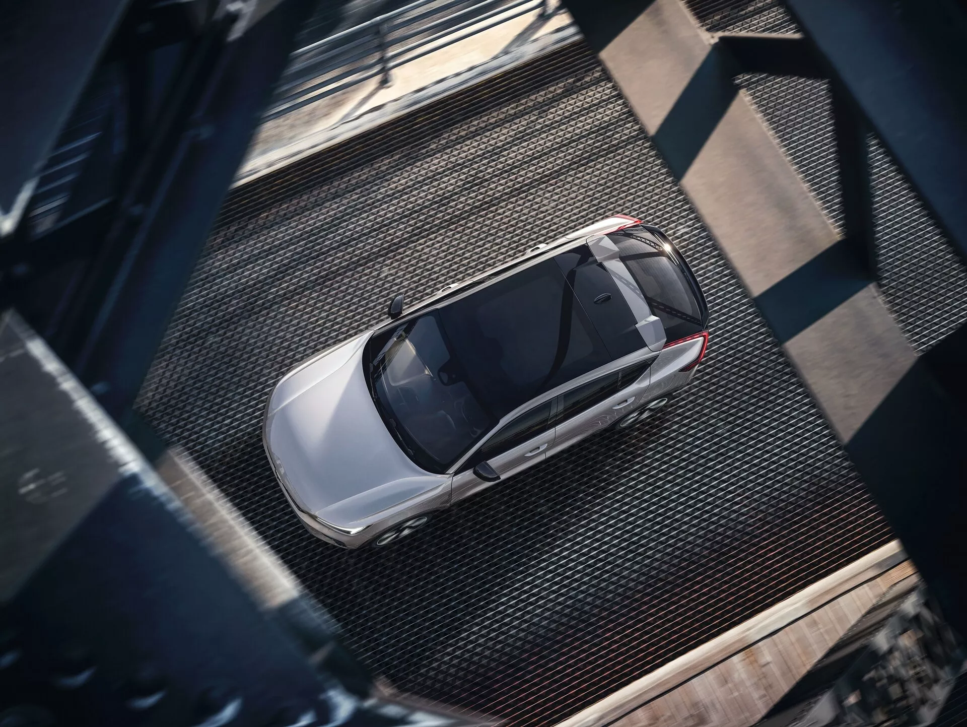 Vue aérienne d'une Volvo C40 2021 de première génération, capturée d'une perspective aérienne. L'image révèle la vue complète de la berline moderne élégante, avec une finition métallique et brillante de couleur claire, probablement argentée ou gris clair. Les caractéristiques visibles incluent le toit, le capot, le coffre, les fenêtres et des parties des portes, avec un toit ouvrant proéminent suggérant une ventilation avancée. La voiture est stationnée sur une surface texturée de style industriel avec des textures métalliques en grille et des poutres rivetées sombres, projetant des ombres suggérant un éclairage latéral probablement le matin ou en fin d'après-midi.