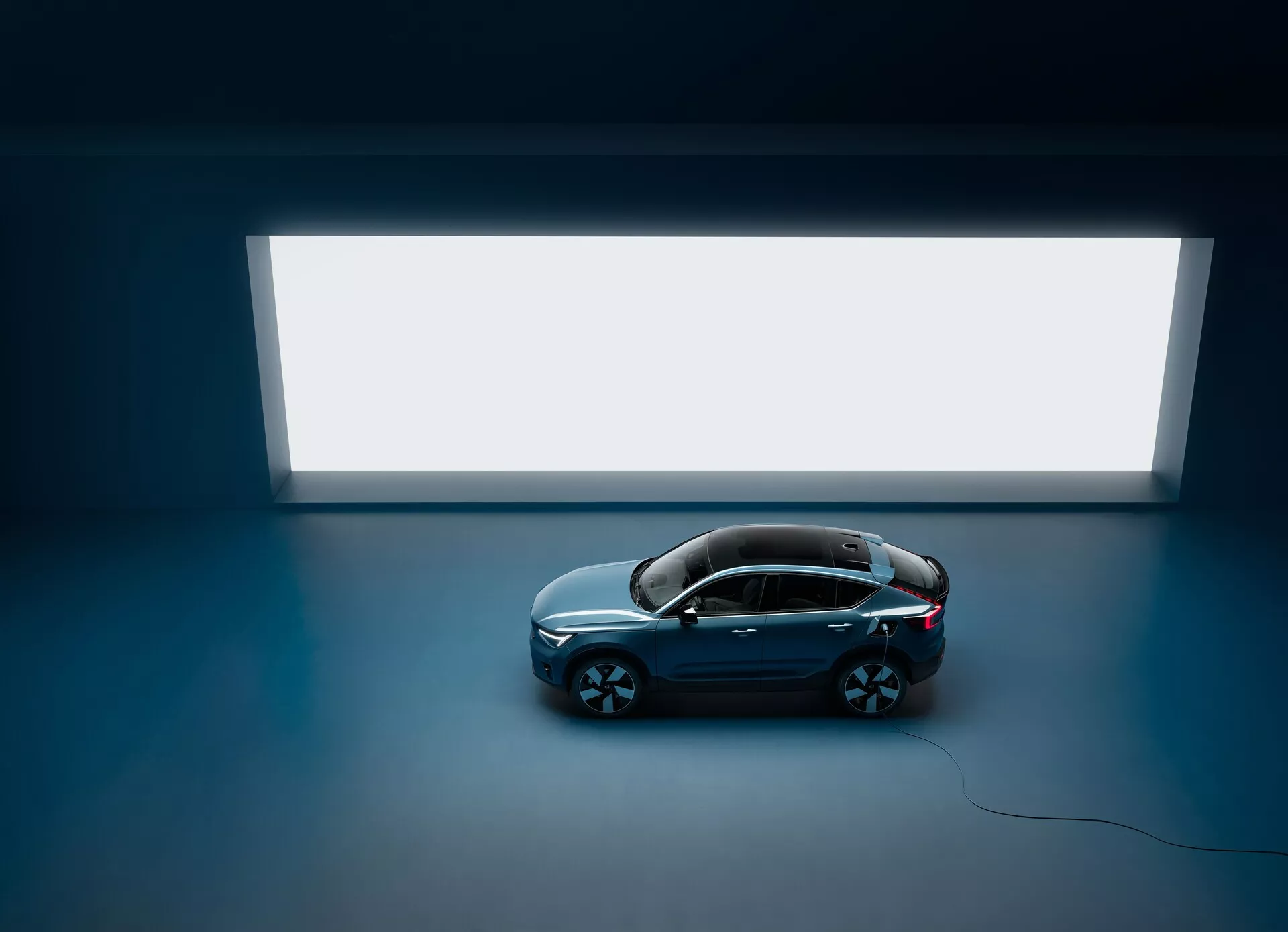 Vue en plongée d'une Volvo C40 2021 berline avec quatre portes et toit panoramique, vue de dessus contre un sol sombre avec une grande source de lumière rectangulaire lumineuse en arrière-plan, présentant un design propre et minimaliste avec une carrosserie de couleur foncée et une finition brillante et de grandes roues en alliage à multiples rayons, soulignant son esthétique luxueuse et moderne.