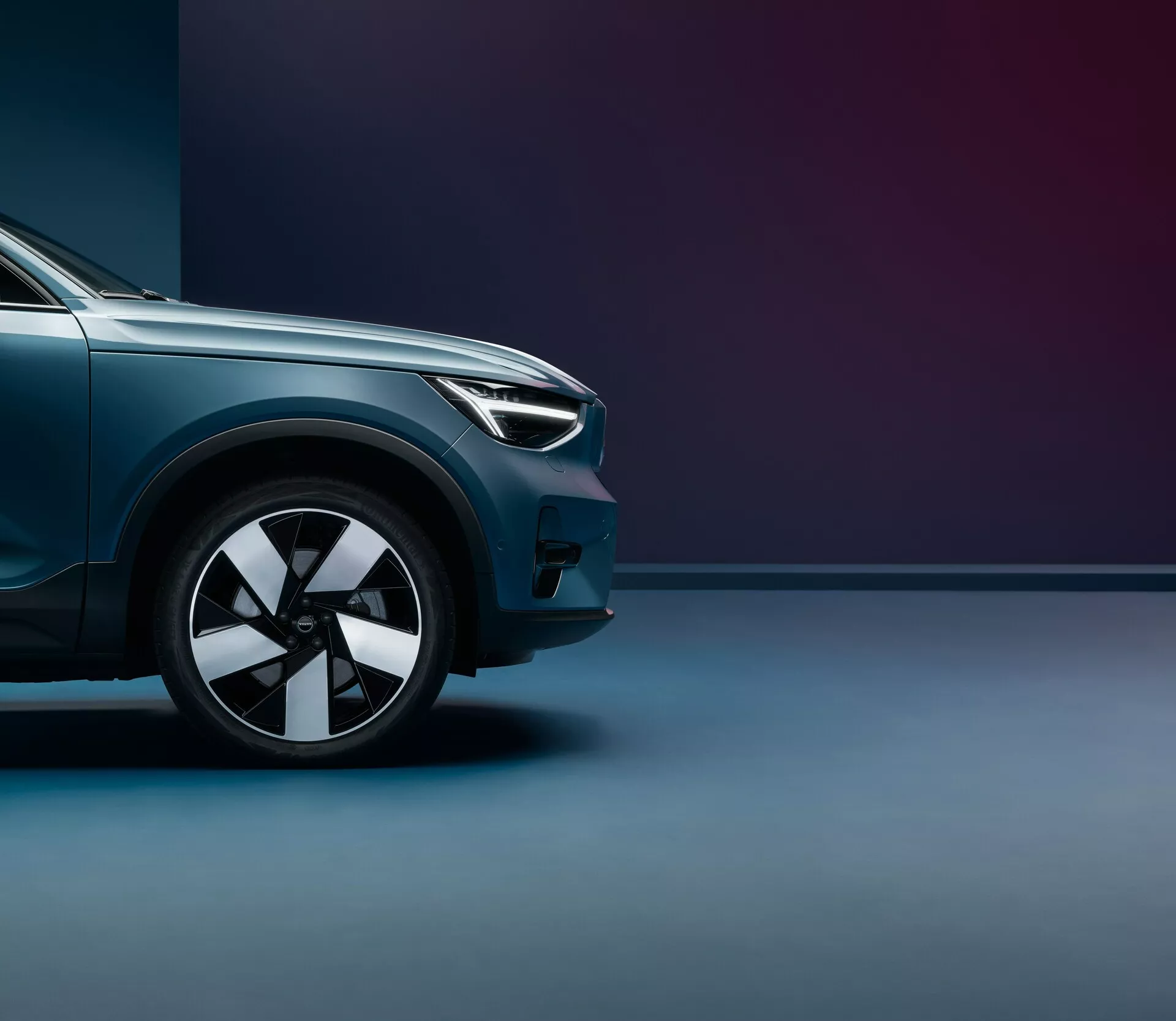 Prise de vue en contre-plongée de la Volvo C40 2021 (1ère génération), se concentrant sur le quart avant, de la roue avant au point central du capot. Sont mis en avant le design moderne des phares, la roue en alliage à plusieurs branches, la peinture métallique brillante et les lignes aérodynamiques. Le fond bicolore avec un éclairage doux accentue la finition premium et la posture imposante du véhicule.