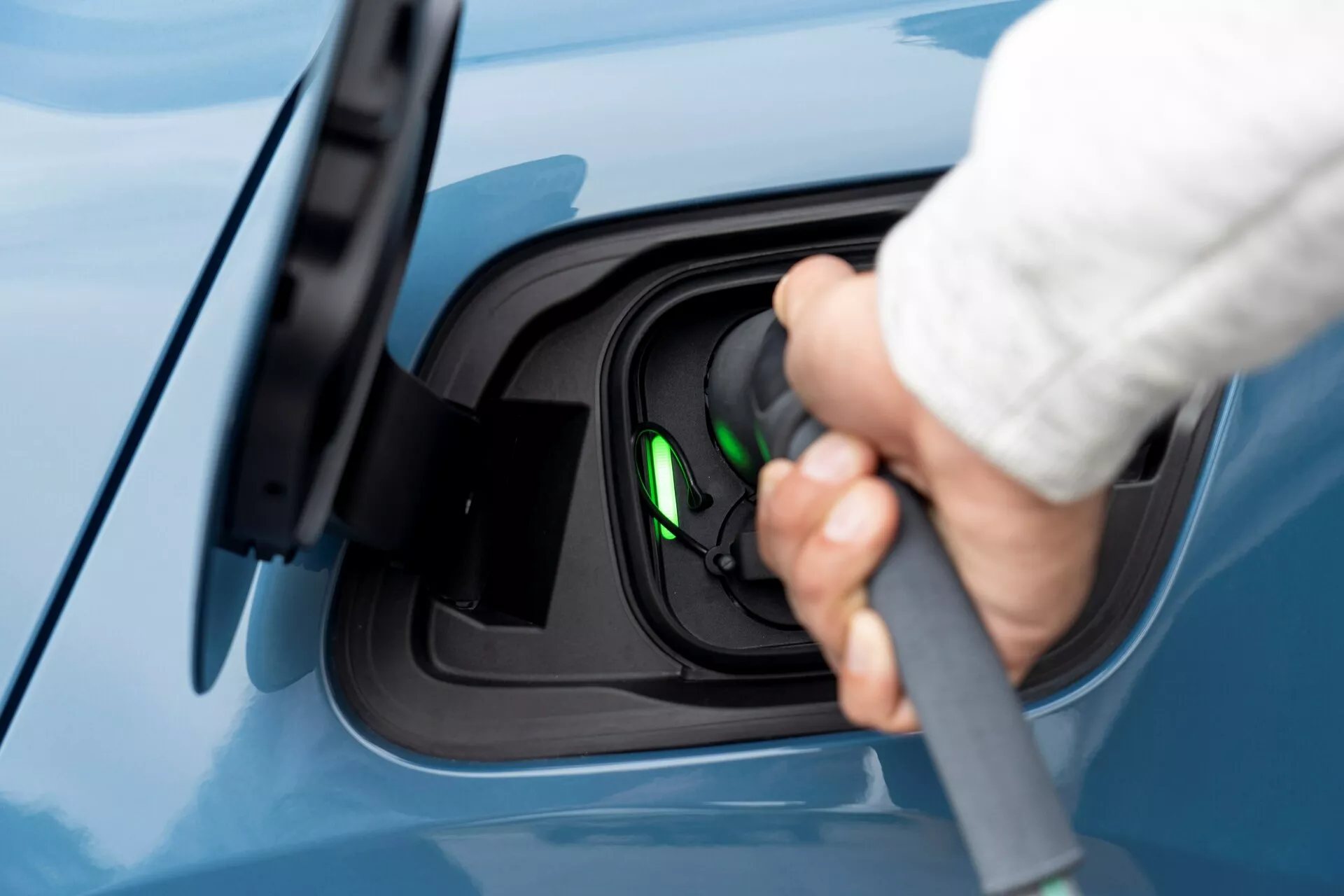Vue latérale rapprochée de la zone de port de charge d'un véhicule électrique Volvo C40 2021 (première génération), avec une main humaine connectant un câble de charge au port. L'image se concentre sur le processus de charge, soulignant le port de charge ouvert avec un connecteur de type 2 probablement utilisé. Une lumière verte à l'intérieur de la prise indique que le véhicule est prêt à être chargé ou est en cours de charge. La vue est légèrement surélevée, montrant une finition de peinture bleue impeccable sur la voiture, soulignant l'aspect propre et moderne du véhicule électrique.