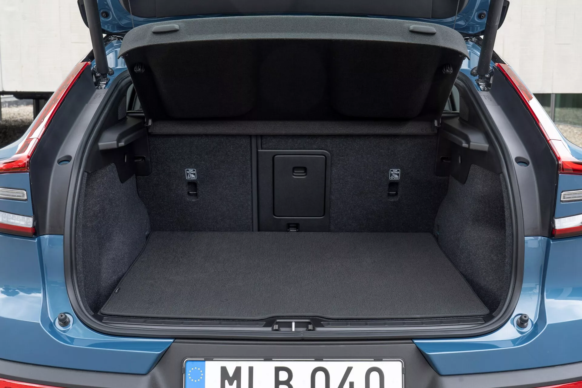 Espace de chargement arrière d'une voiture moderne, montrant le coffre de la Volvo C40 2021 (1ère génération) d'une perspective arrière, légèrement en dessous de la taille. Le couvercle du coffre est ouvert, mettant en évidence le rembourrage standard de la doublure intérieure. Le coffre est vide, affichant l'intérieur en moquette, les zones latérales encastrées et une poignée au milieu du plancher pour accéder à un compartiment de rangement. Aucun crochet de chargement ou point d'ancrage n'est visible. La plaque d'immatriculation de style européen suggère une origine européenne.