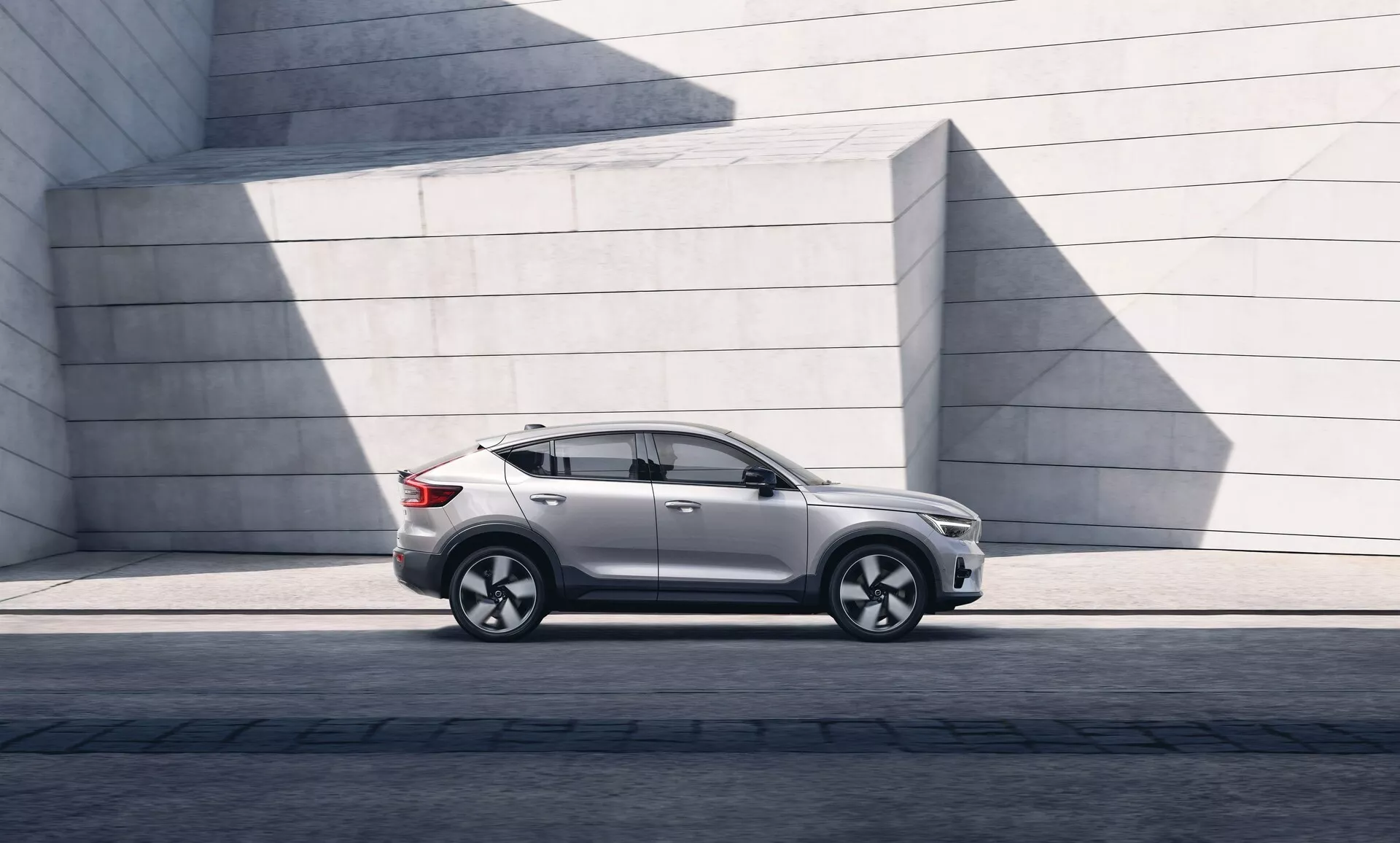 Image d'un SUV moderne Volvo C40 2021 garé parallèlement, montrant le profil latéral gauche du phare avant gauche au feu arrière gauche. L'angle met en avant la garde au sol et la posture sur une surface pavée. La lumière du soleil crée des ombres sur la finition de peinture brillante. Le SUV présente un design élégant avec une ligne de toit doucement inclinée, des contours de portes distincts, des fenêtres teintées et des rétroviseurs contemporains. Les roues en alliage avec un design multi-rayons et des pneus à profil bas indiquent performance et luxe. Le bas de caisse, les moulures d'arches de roues et une ligne de caractère à travers les poignées de portes sont visibles, suggérant la robustesse. L'absence de visible échappement laisse penser à une possible motorisation électrique ou hybride. Les structures en béton minimalistes de l'arrière-plan contrastent avec les courbes raffinées de la voiture, accentuant son attrait urbain et design.