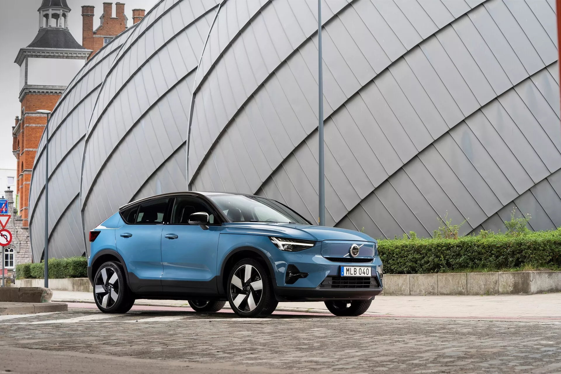 Un SUV Volvo C40 2021 moderne bleu garé sur une zone pavée urbaine avec une structure angulaire au look métallique en arrière-plan. La prise de vue en contre-plongée depuis le côté avant gauche montre la calandre avant du véhicule, les phares à LED, le profil aérodynamique, les jantes en alliage à plusieurs branches, les rétroviseurs avec clignotants intégrés, la finition de peinture mate ou satinée, les vitres teintées, la plaque d'immatriculation avant et les lignes sculptées du corps.