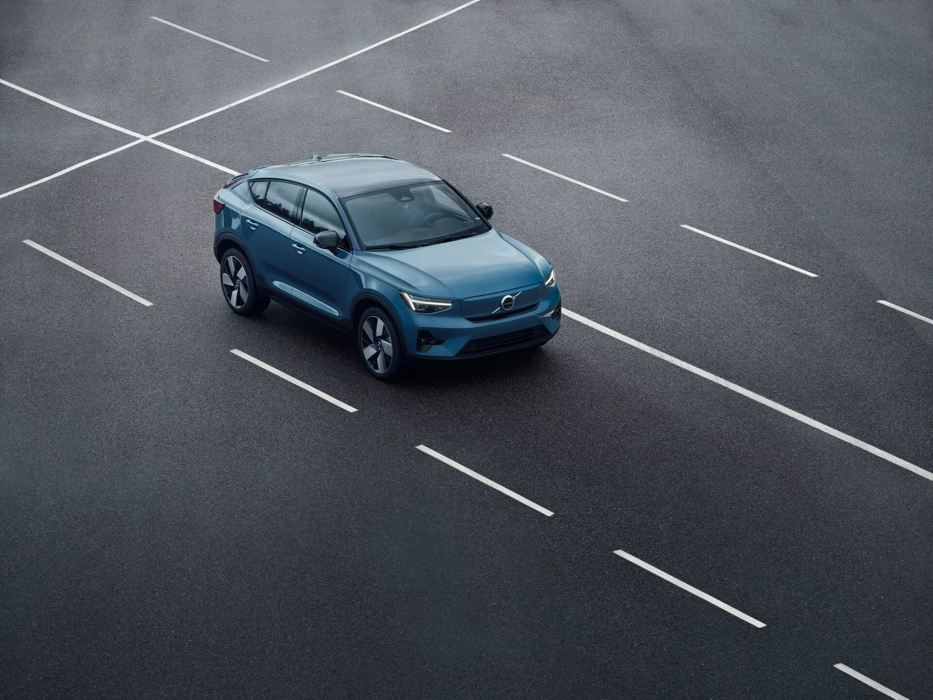 Vue aérienne d'une Volvo C40 2021 (1ère génération) dans un parking, positionnée en diagonale sur plusieurs places. L'image capture les profils supérieur et latéral de la voiture, mettant en évidence le toit, le capot, les rétroviseurs latéraux, les roues et la partie supérieure de la grille. Le design élégant et moderne, le toit ouvrant éventuel, les barres de toit et les phares à LED suggèrent des avancées technologiques.