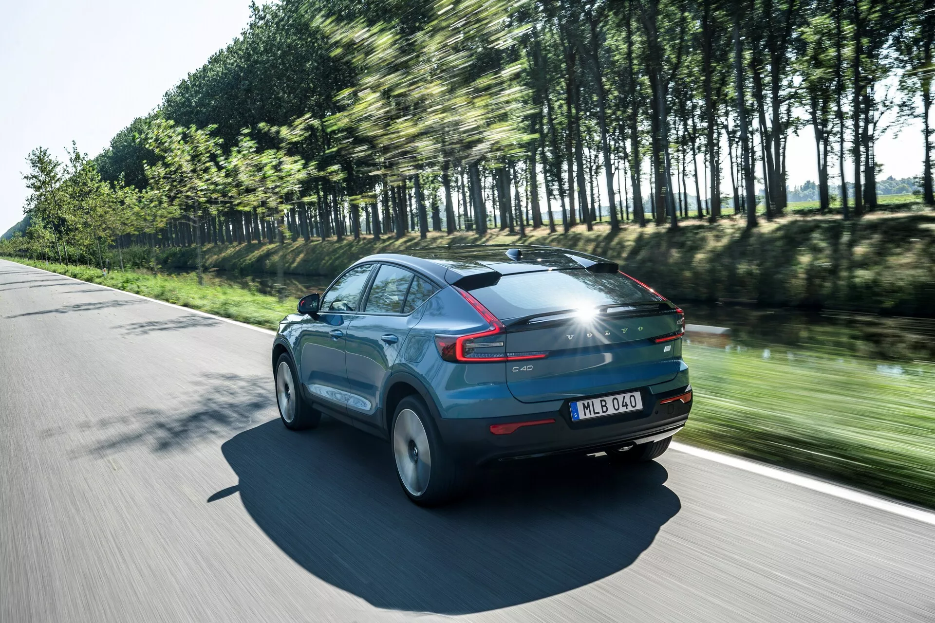 Prise de vue dynamique de la Volvo C40 2021 (1ère génération) en mouvement par une journée ensoleillée, vue arrière trois quarts mettant en avant le profil aérodynamique du véhicule. La voiture, peinte en bleu brillant, présente des feux arrière LED distinctifs, des roues floues en mouvement et des arbres reflétant sa vitesse. Des détails notables incluent un toit panoramique, un aileron arrière et une plaque d'immatriculation sur le hayon, soulignant des éléments de design moderne et une technologie d'éclairage avancée.