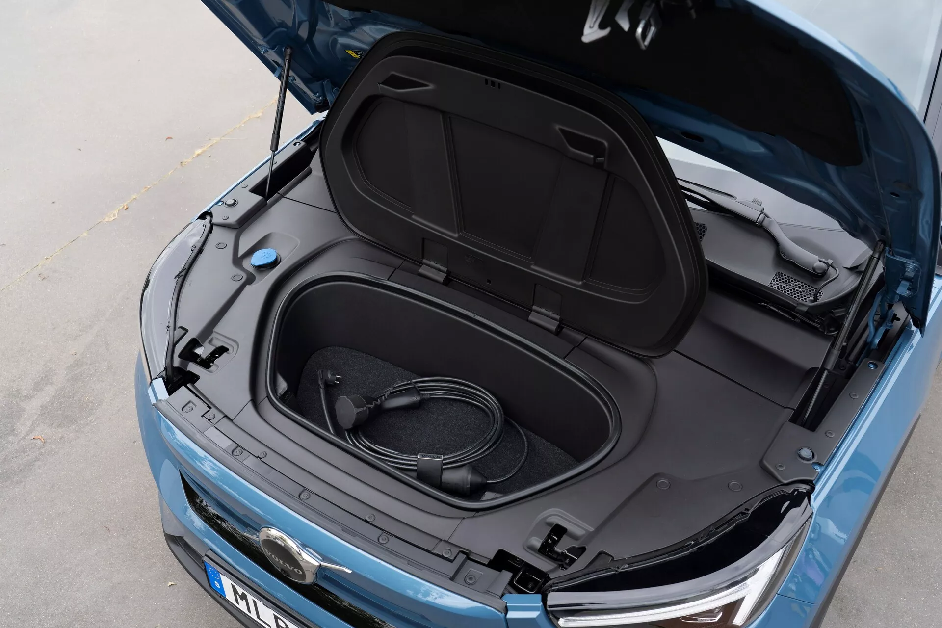 Vue rapprochée en plongée angulaire de la Volvo C40 2021 (1ère génération) avec son frunk ouvert, révélant un espace de rangement vide et un câble de recharge enroulé, indiquant qu'il s'agit d'un véhicule électrique. Peinture bleu métallisé, intérieur du capot avant noir avec des autocollants informatifs, vue partielle du logo du fabricant, phares élégants et grille avant visibles, suggérant un VE avec technologie LED moderne et esthétique aérodynamique.