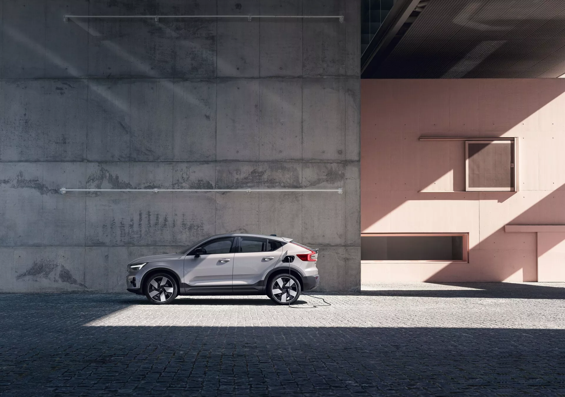 Profil latéral du SUV moderne Volvo C40 2021 (1ère Génération) contre un arrière-plan urbain avec des murs en béton et de couleur pêche. La voiture présente une peinture blanche réfléchissante, des jantes en alliage élégantes, des vitres teintées, des barres de toit argentées et des clignotants intégrés dans les rétroviseurs. La photo met en valeur les lignes et courbes du design de la voiture sous la lumière naturelle du jour, ajoutant une touche de luxe et de sophistication.