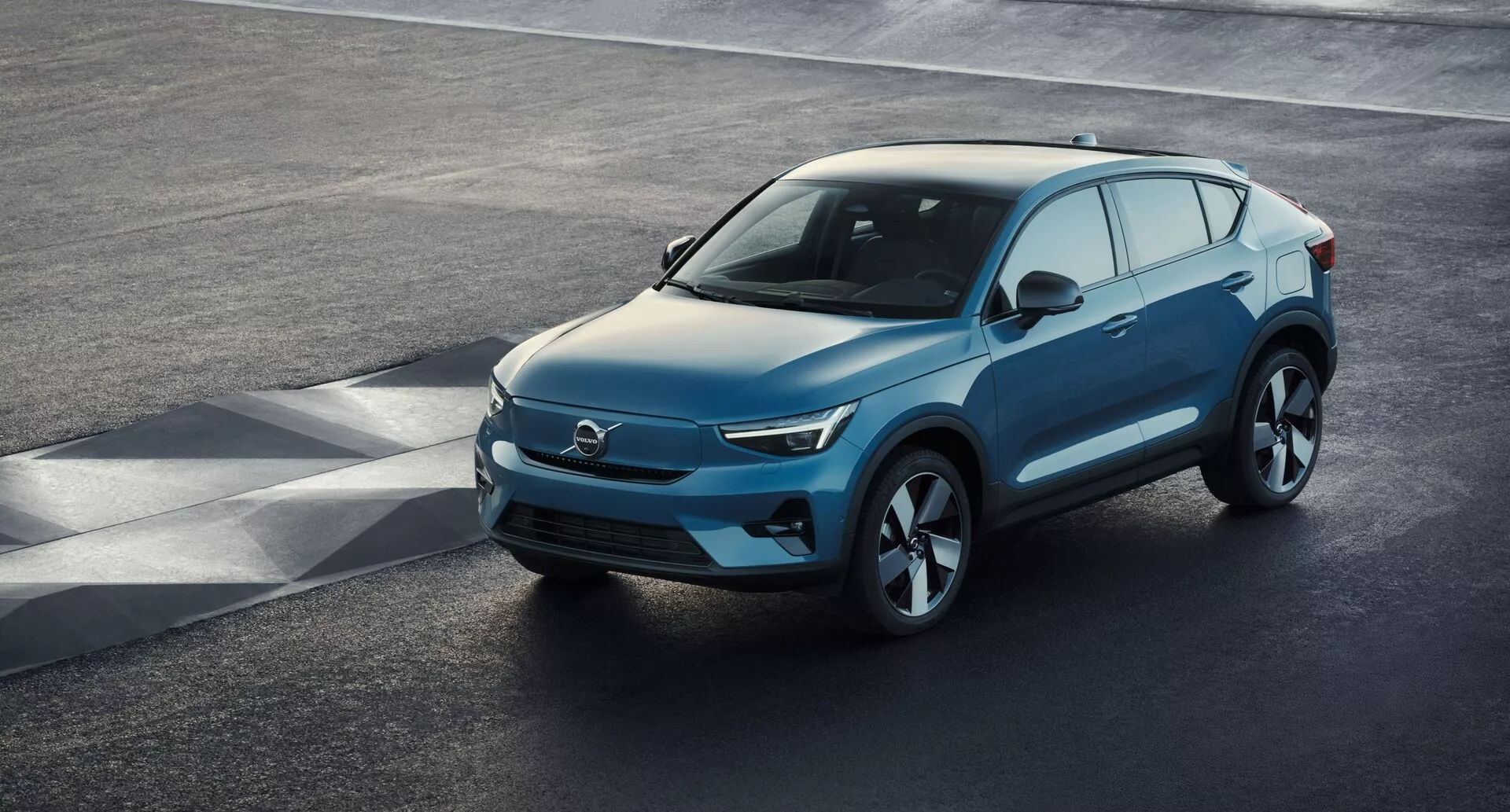 Vue en angle élevé d'un SUV moderne Volvo C40 2021 (1re génération) garé sur une surface de goudron, montrant une perspective frontale trois-quarts élevée. La zone avant élégante, probablement caractéristique d'un véhicule électrique avec des prises d'air minimales, se combine avec des phares à LED nets, un capot lisse en pente et un profil latéral profilé avec de grandes roues en alliage. Rétroviseurs latéraux avec signal de clignotant intégré, large espace de vitres avec possible teinte de confidentialité et une ligne de toit légèrement inclinée avec barres de toit pour l'utilité sont des caractéristiques notables. La vue claire capture le design luxueux du véhicule, l'aérodynamique avancée et les éléments de technologie moderne.