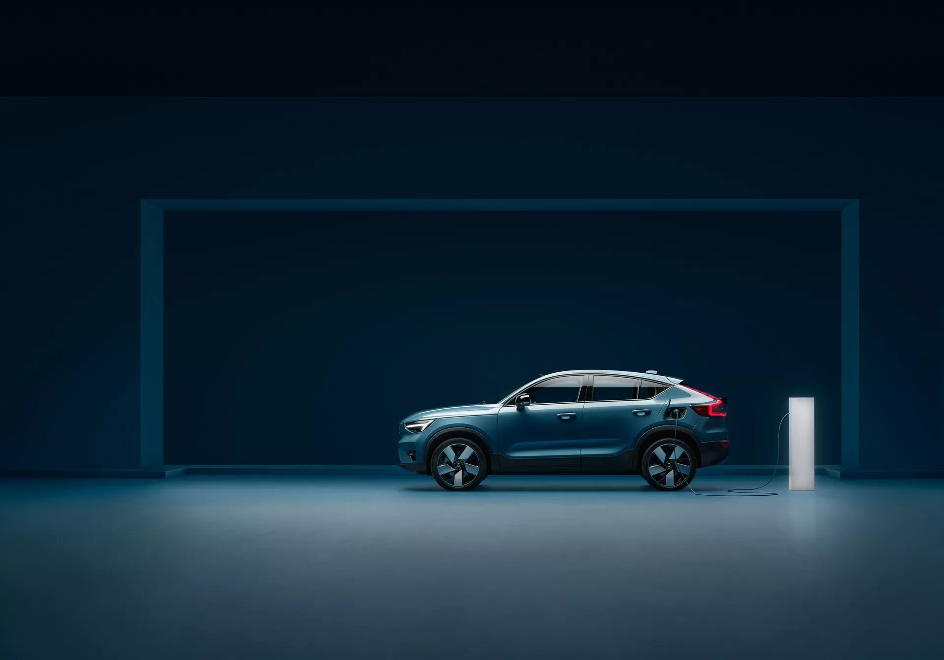 Vue de profil latéral d'une voiture moderne Volvo C40 2021 (première génération) dans un studio avec un sol réfléchissant, montrant un design élégant, une peinture métallique bleu foncé, une ligne de toit inclinée, de grandes jantes en alliage à rayons multiples, des lignes profilées, une construction robuste, des rétroviseurs latéraux intégrés avec clignotants, des feux arrière à LED et une prise de recharge électrique.