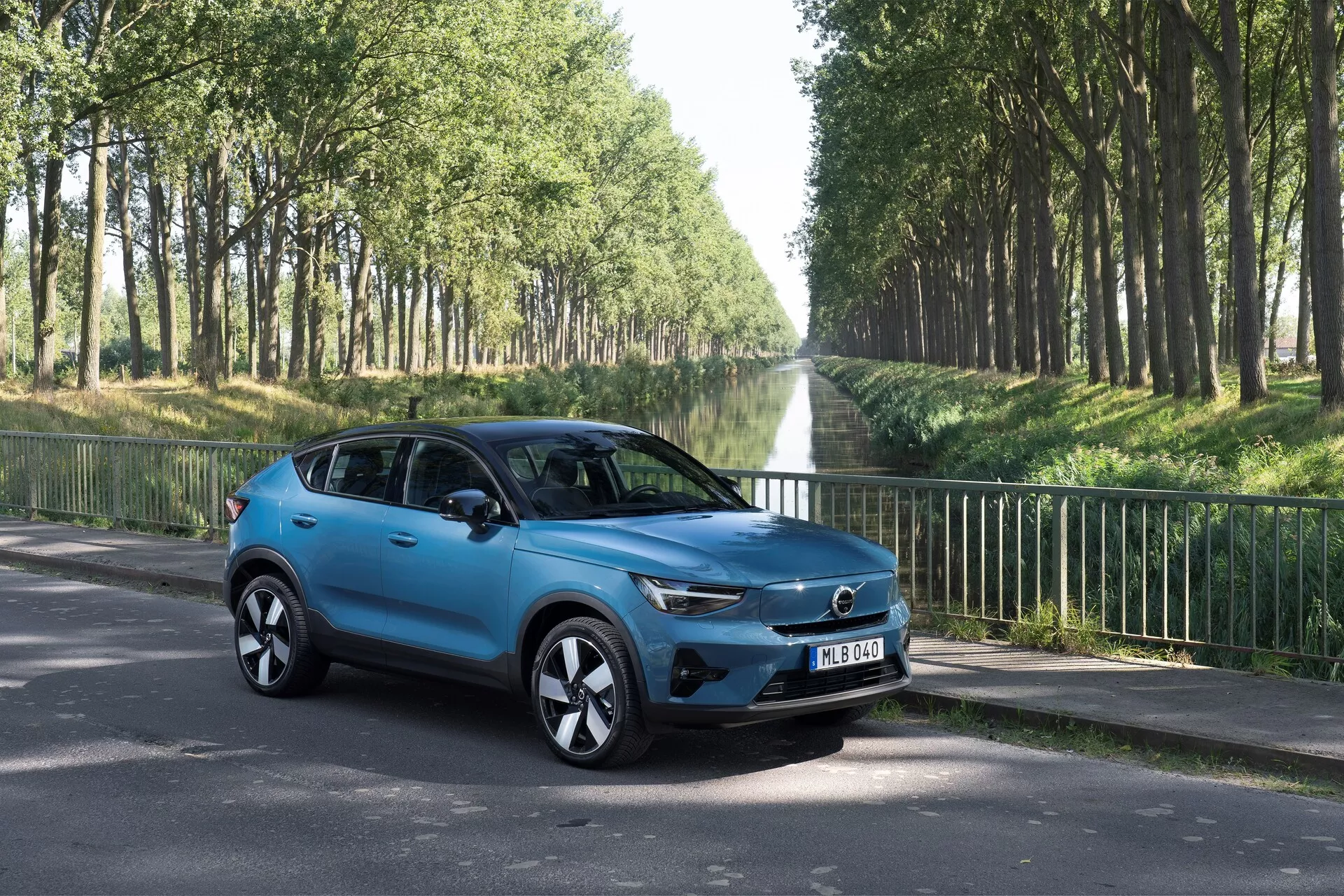 Volvo C40 2021 (Première Génération) SUV compact vu de trois quarts avant gauche sur une route en asphalte. Présente des caractéristiques de design moderne comme la calandre proéminente avec badge central, des phares LED élégants, des jantes en alliage stylisées, des rétroviseurs latéraux avec clignotants intégrés, une carrosserie sculptée aérodynamique avec une finition de peinture bleue brillante, des fenêtres teintées et quatre portes. Environnement extérieur avec des arbres bordant la route, axé sur les caractéristiques esthétiques et technologiques du véhicule.