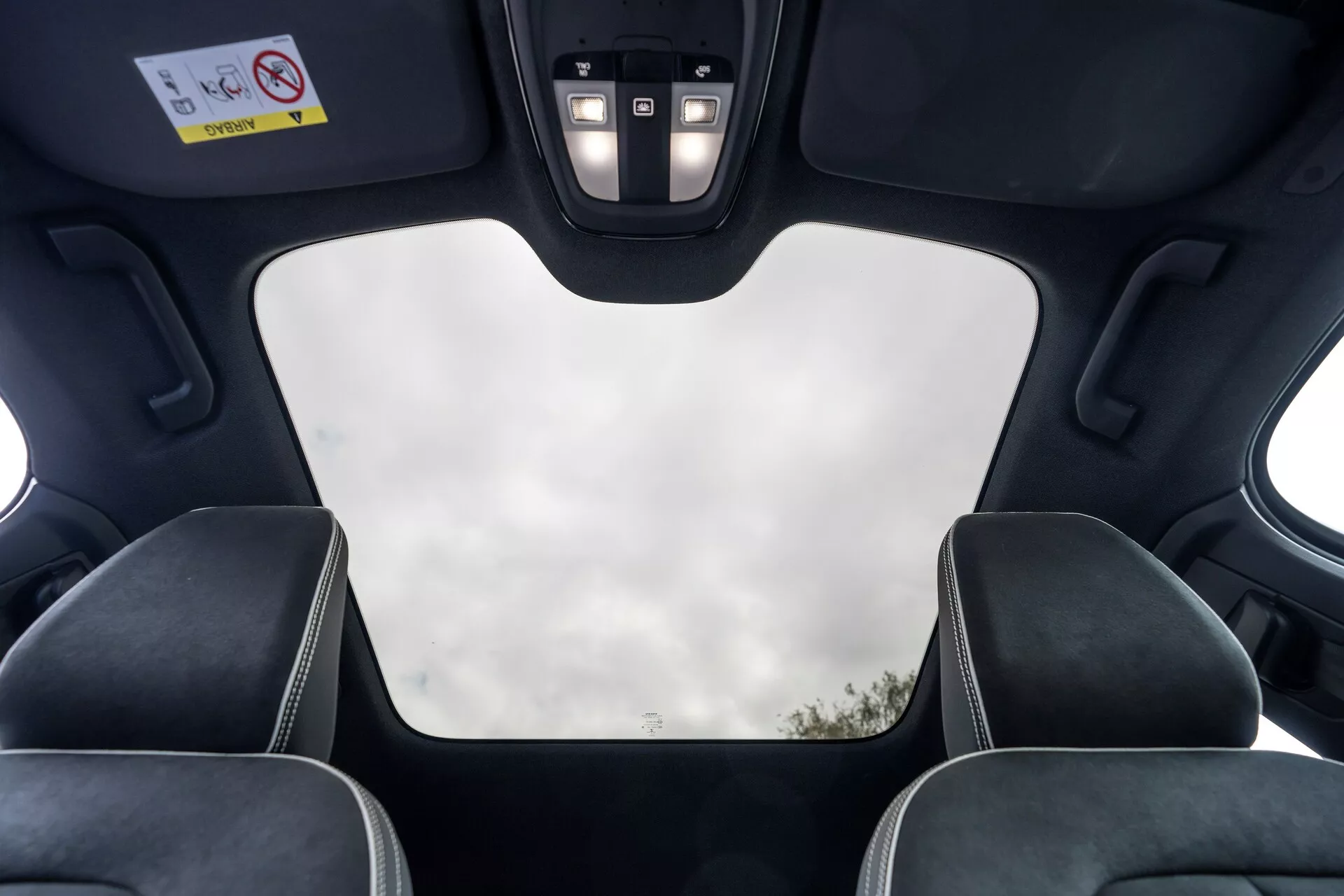 Vue intérieure de la Volvo C40 2021 (1ère génération) depuis la perspective du passager arrière, mettant en évidence des éléments de toit tels qu'un toit panoramique fermé avec vue dégagée du ciel, un revêtement intérieur noir, des découpes pour le mécanisme de toit, des lumières LED et un panneau de contrôle avec boutons pour diverses fonctions. L'image met également en avant les appuie-têtes des sièges avant avec coutures contrastantes, soulignant le design moderne et luxueux du véhicule et ses caractéristiques haut de gamme.
