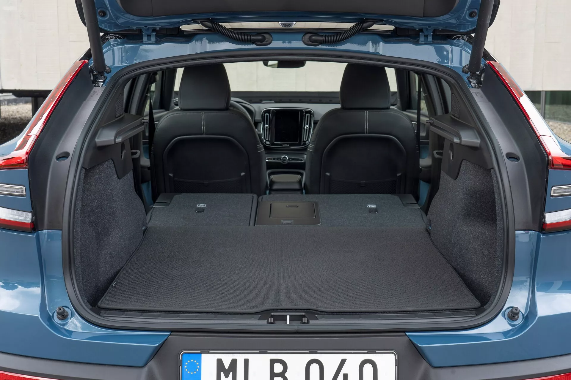 Intérieur arrière de la Volvo C40 2021 (1ère génération) avec le coffre ouvert, montrant l'espace de chargement vide avec une doublure en tapis durable, une surface plate et sans intrusions visibles. La partie supérieure des sièges arrière en tissu foncé est visible, avec une vue partielle des éléments de la cabine avant tels que la console centrale et le système d'infodivertissement. Le couvercle du coffre est levé, révélant les bords en plastique/composite.