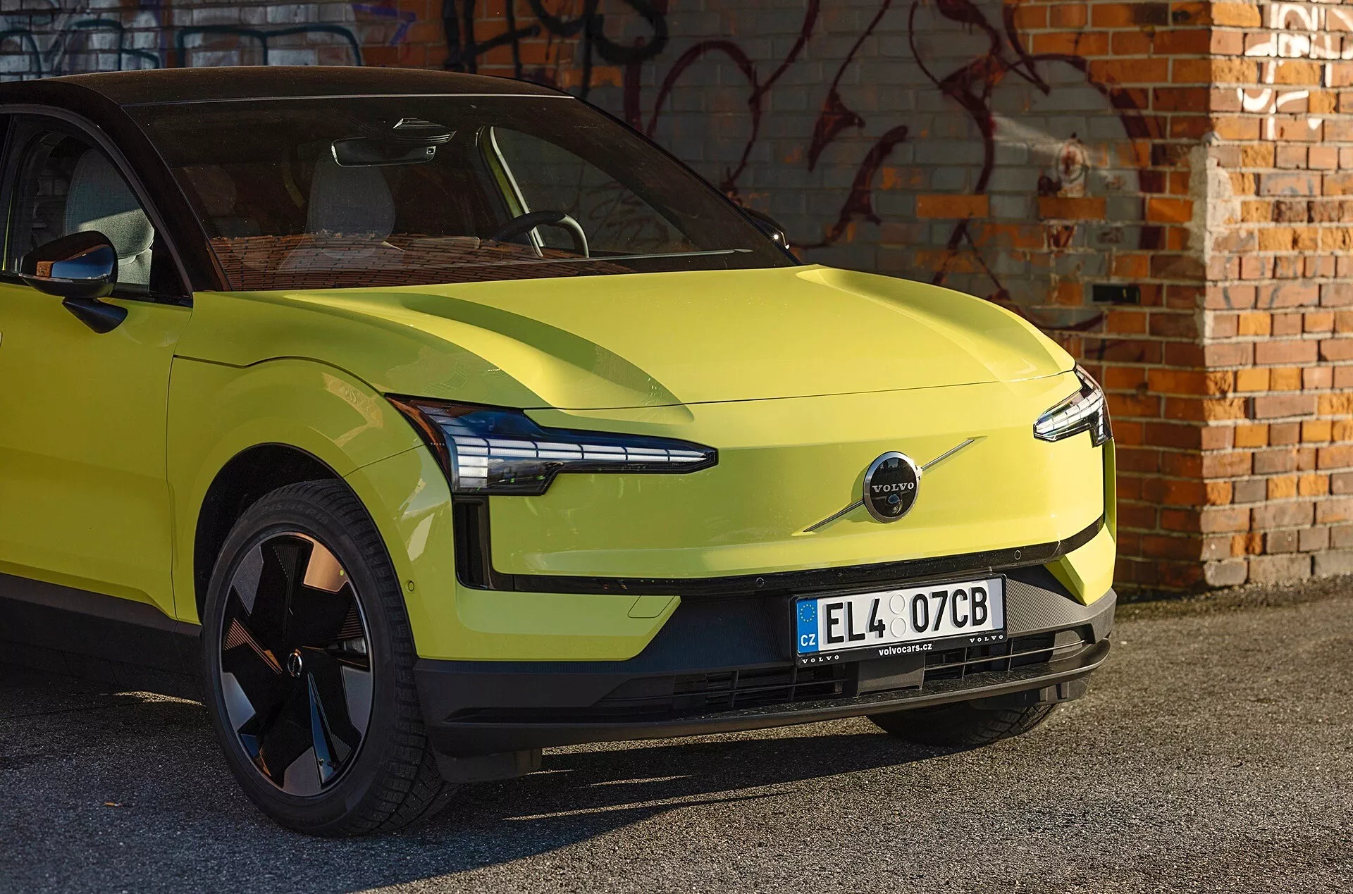 Voiture électrique Volvo EX30 vert lime de 2024 garée devant un mur en briques avec graffiti, présentant un design aérodynamique, des phares LED modernes et l'absence de calandre avant