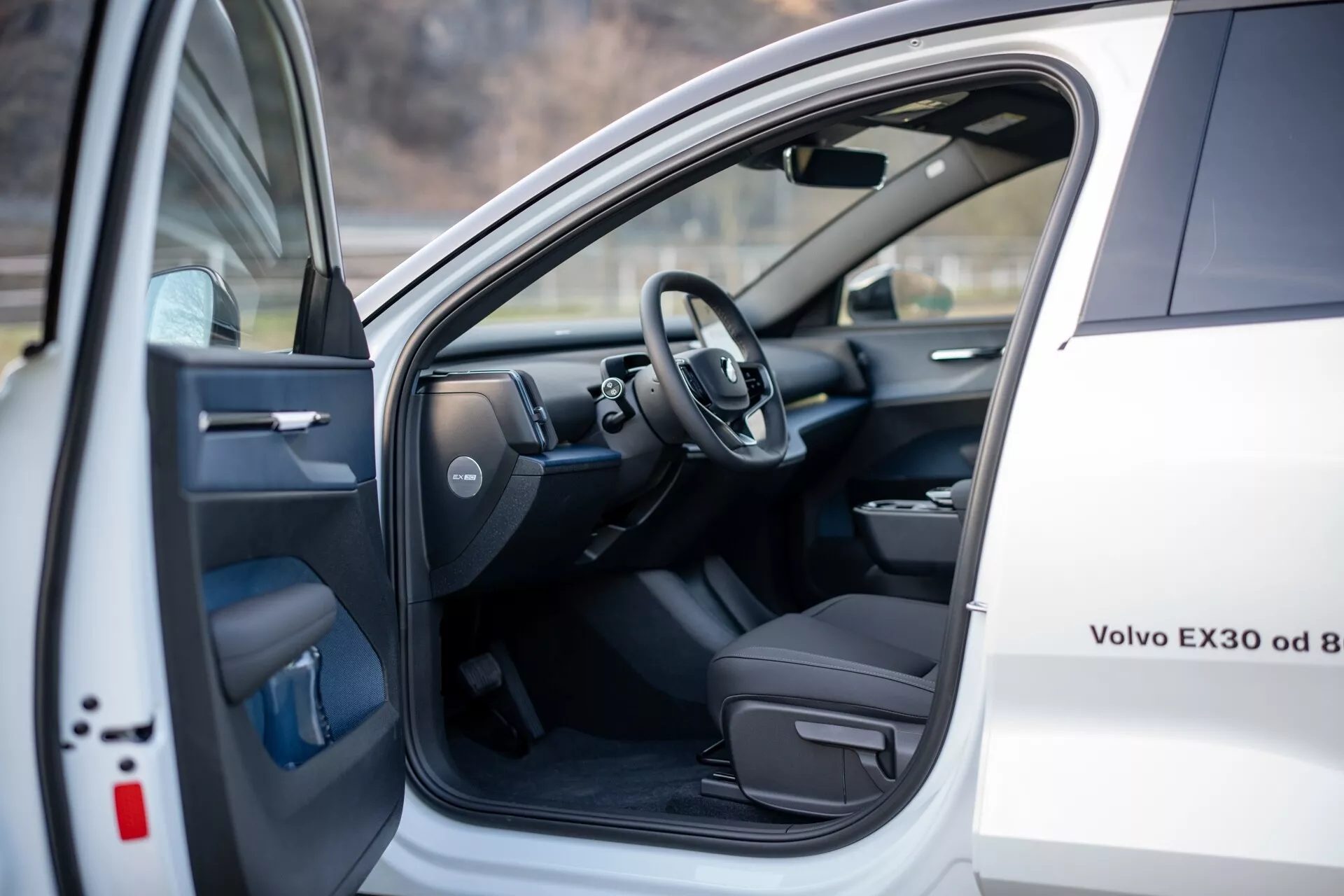 Vue intérieure d'une Volvo EX30 blanche 2024, montrant le siège conducteur et la zone cockpit avec la porte avant gauche ouverte comportant des commandes électriques, et un aperçu du volant et d'une partie du tableau de bord