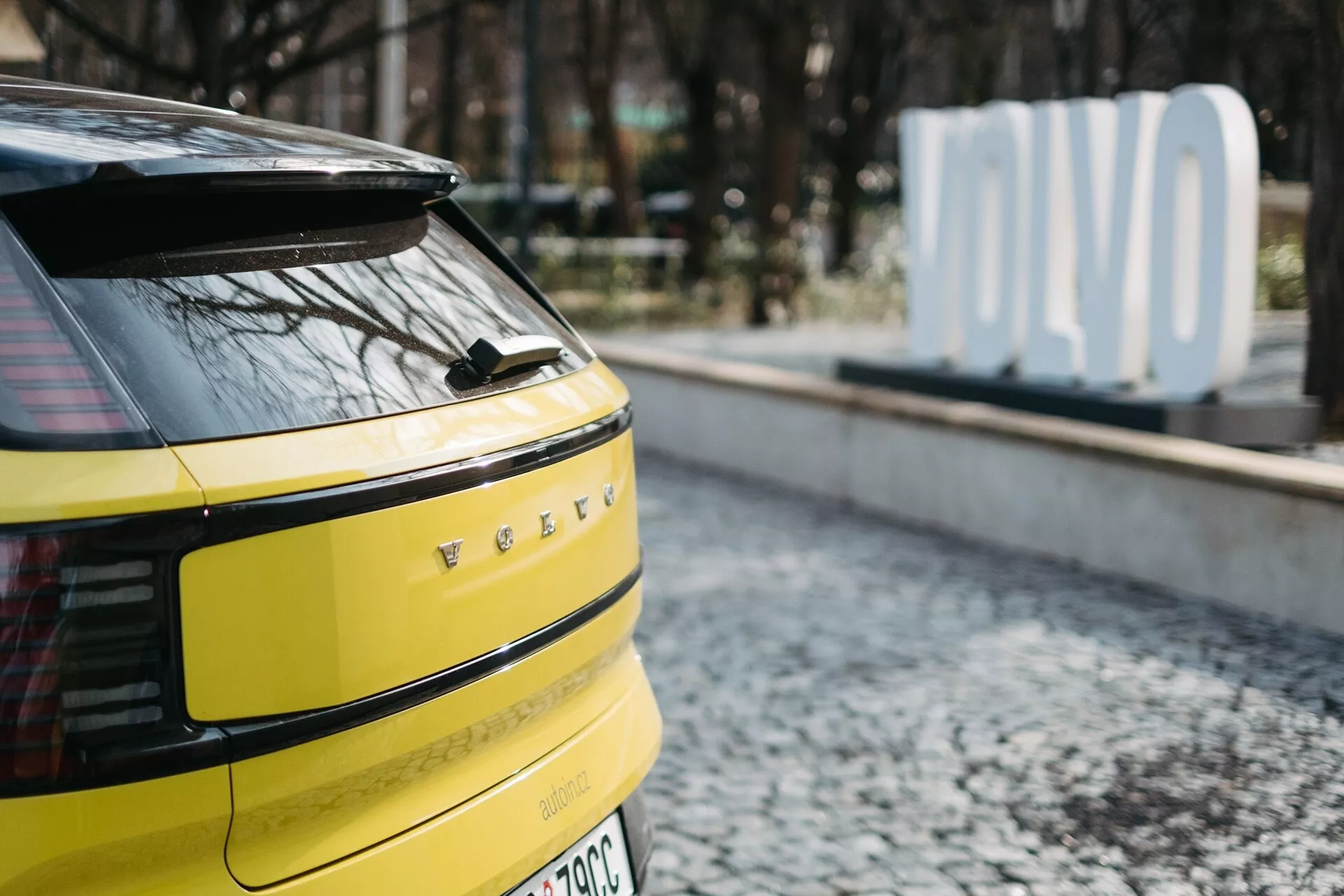 Gros plan sur la section arrière d'une Volvo EX30 2024 en jaune vif, avec feux arrière LED horizontaux, caméra ou capteurs arrière potentiels, essuie-glace arrière unique, badge de véhicule électrique ou hybride et antenne aileron de requin, garée sur une surface pavée avec un panneau 'OLVO' flou en arrière-plan