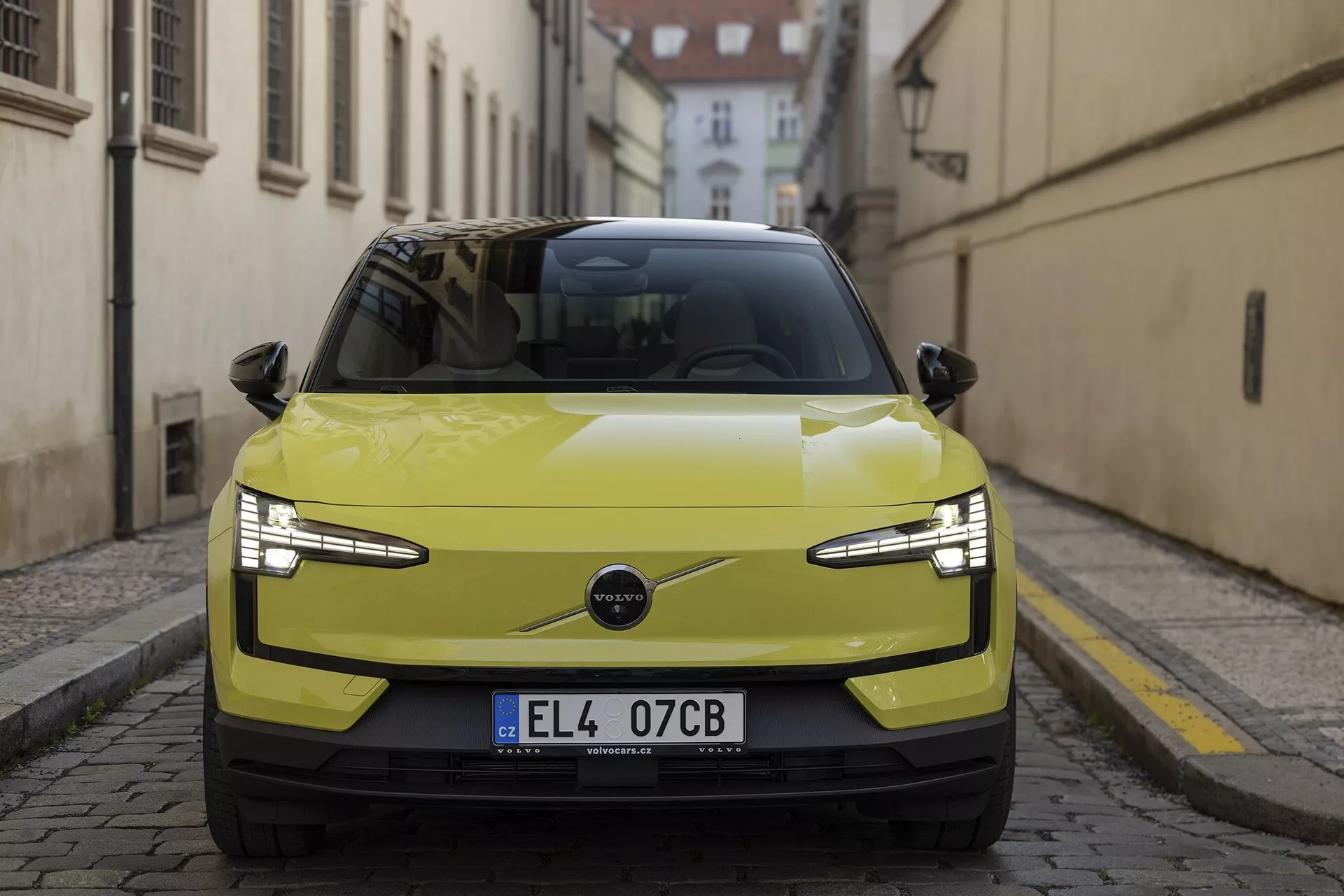 Vue frontale de la Volvo EX30 2024 en jaune citron, mettant en avant ses phares LED élégants, sa calandre minimaliste suggérant une motorisation électrique et ses fonctionnalités technologiques avancées comme les capteurs de stationnement et l'ADAS possible, devant une rue historique européenne