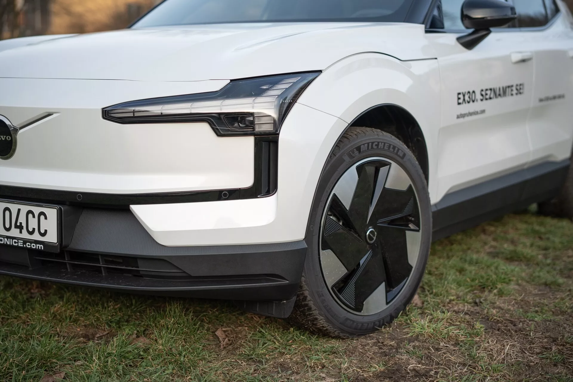 Vue rapprochée du véhicule électrique Volvo EX30 2024 depuis une perspective de trois quarts, mettant en évidence son phare LED, design de roue aérodynamique et pare-choc avant minimaliste sans grille, en blanc avec garniture noire.