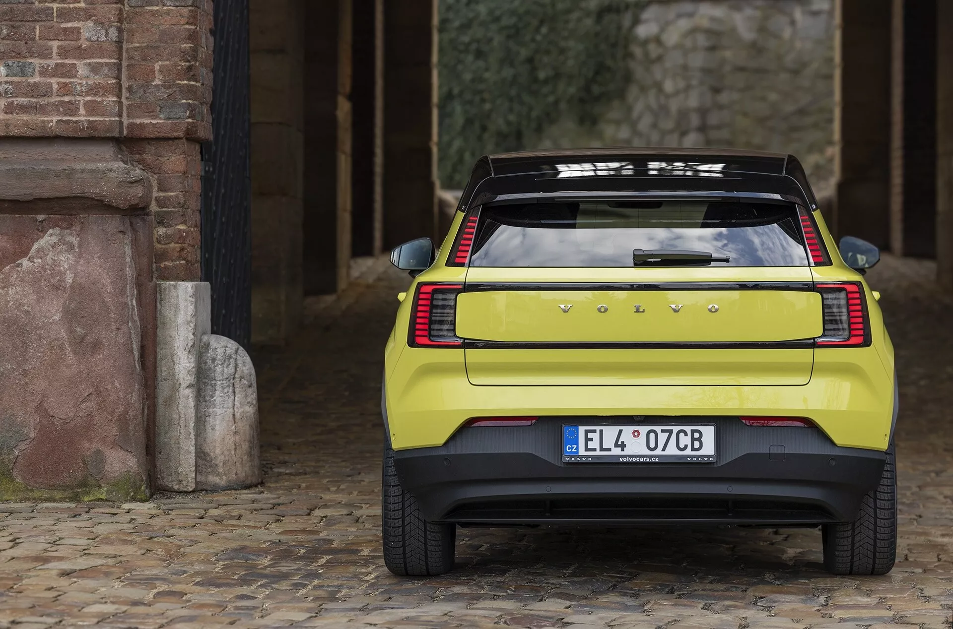 Vue arrière de la Volvo EX30 2024 sur pavés montrant des feux arrière horizontaux élégants, un emblème du fabricant, une plaque d'immatriculation européenne, un grand pare-brise arrière avec dégivreur, un spoiler sportif et un pare-chocs robuste avec capteurs de stationnement