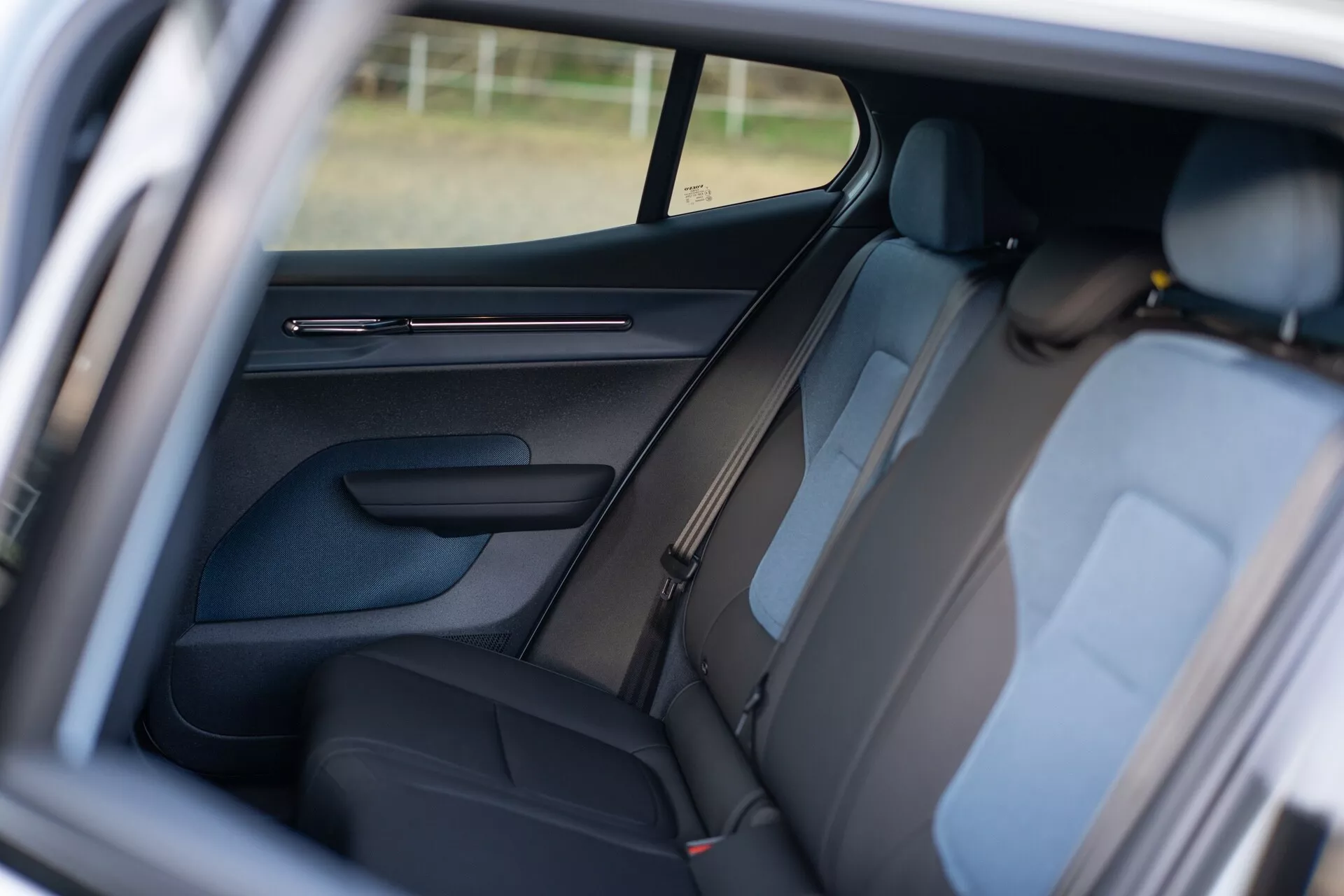 Intérieur du Volvo EX30 2024 montrant la zone des passagers arrière à travers la porte du passager avant ouverte, présentant une sellerie foncée, des appuie-têtes visibles, un panneau de porte détaillé avec commandes, design du montant B, ceintures de sécurité pour les sièges arrière, une fenêtre latérale partiellement visible améliorant l'ambiance intérieure, et un accent sur la disposition des sièges pour le confort et la sécurité des passagers.