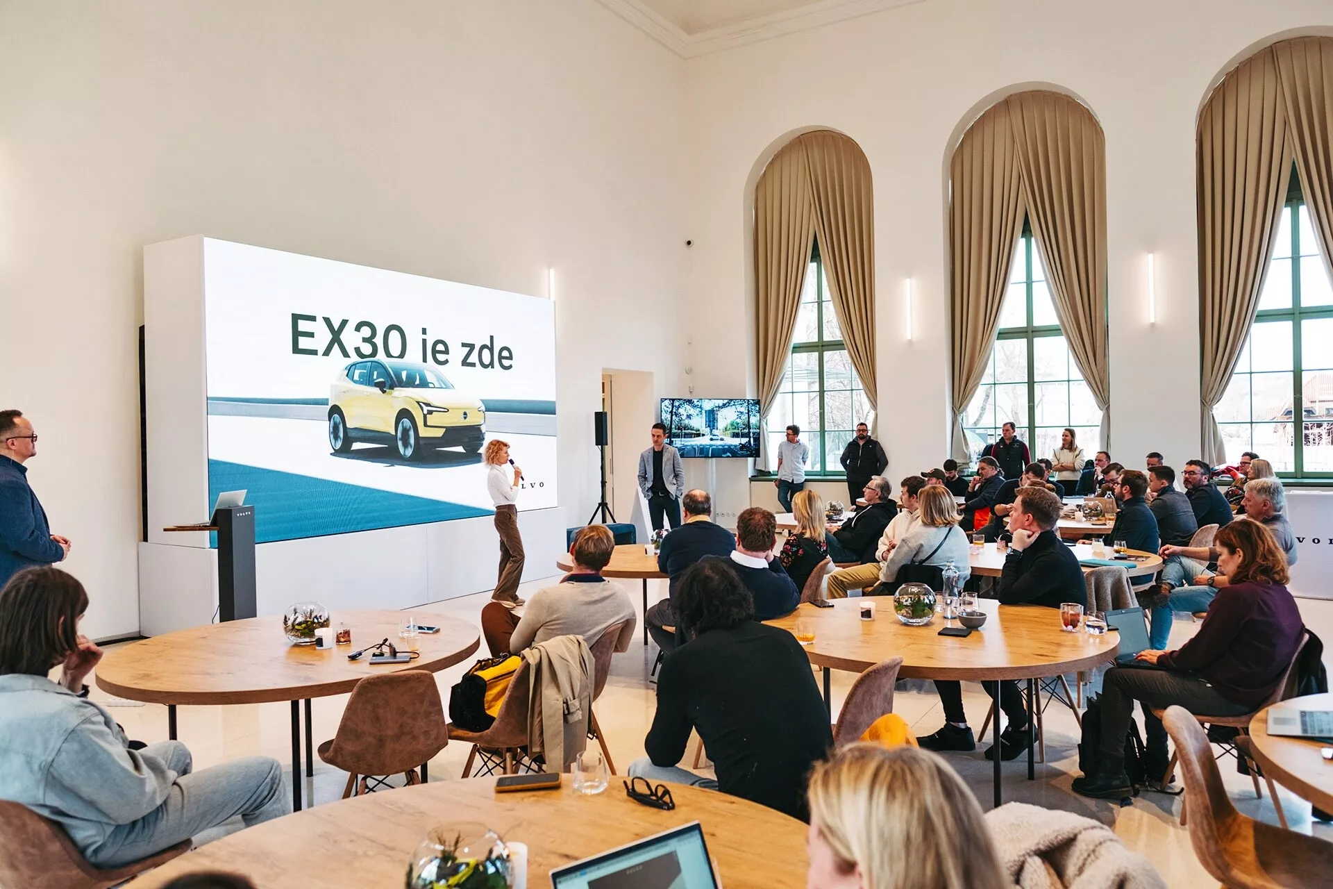 Présentation du véhicule électrique Volvo EX30 2024 dans une salle spacieuse avec de hauts plafonds et de grandes fenêtres voûtées, mettant en vedette un EX30 jaune vu de trois quarts avant sur un grand écran