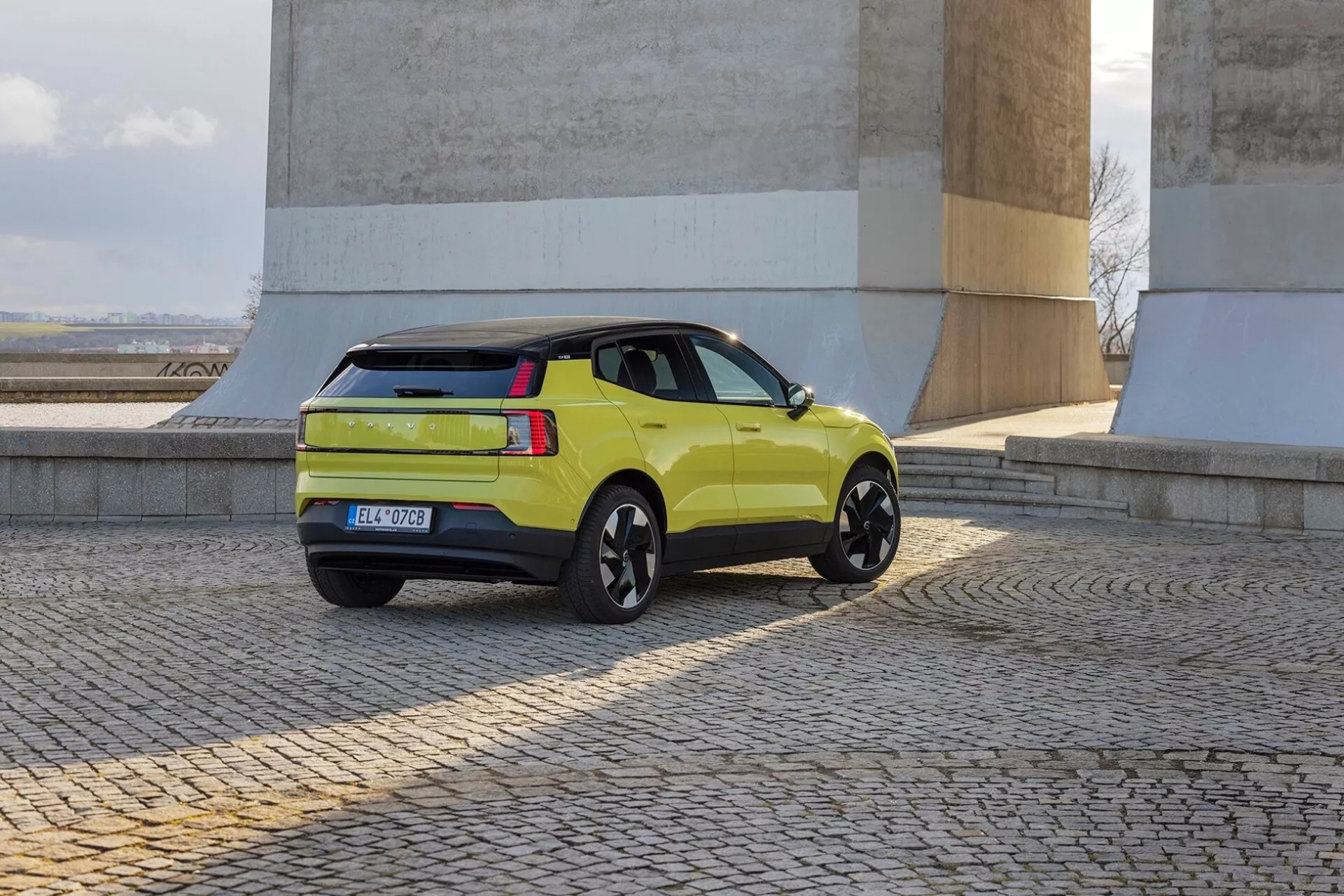 SUV vert lime Volvo EX30 2024 en vue arrière à trois quarts présentant profil latéral, feux arrière, hayon et jantes alliage noires près de murs en pierre sur sol pavé