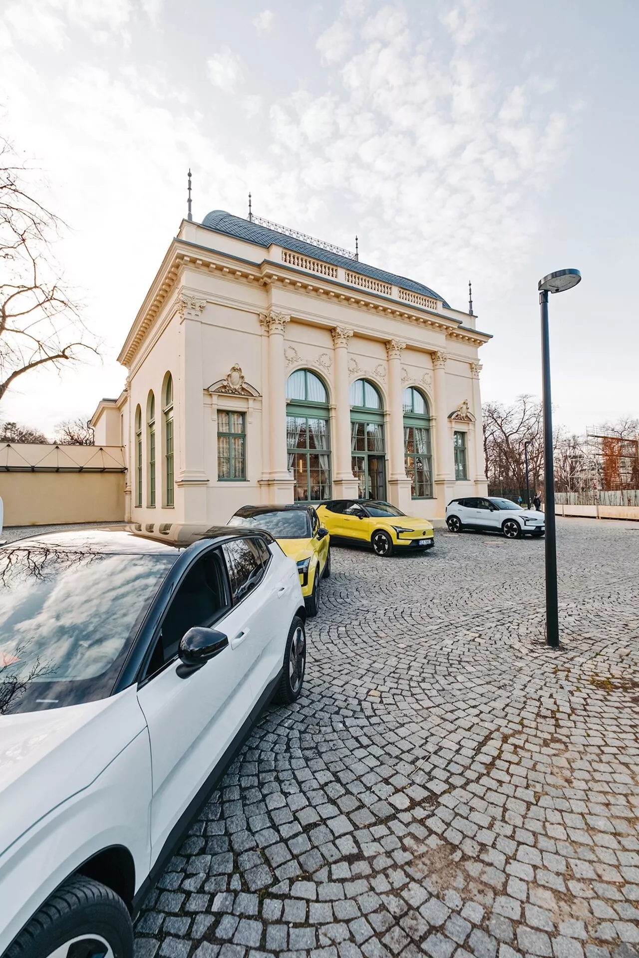 Scène de rue avec trois voitures et un bâtiment élégant en arrière-plan, prise d'un point de vue grand-angle mettant en évidence une berline blanche contemporaine, une voiture de sport jaune vibrante, et une berline ou hatchback blanche à la lumière de fin d'après-midi