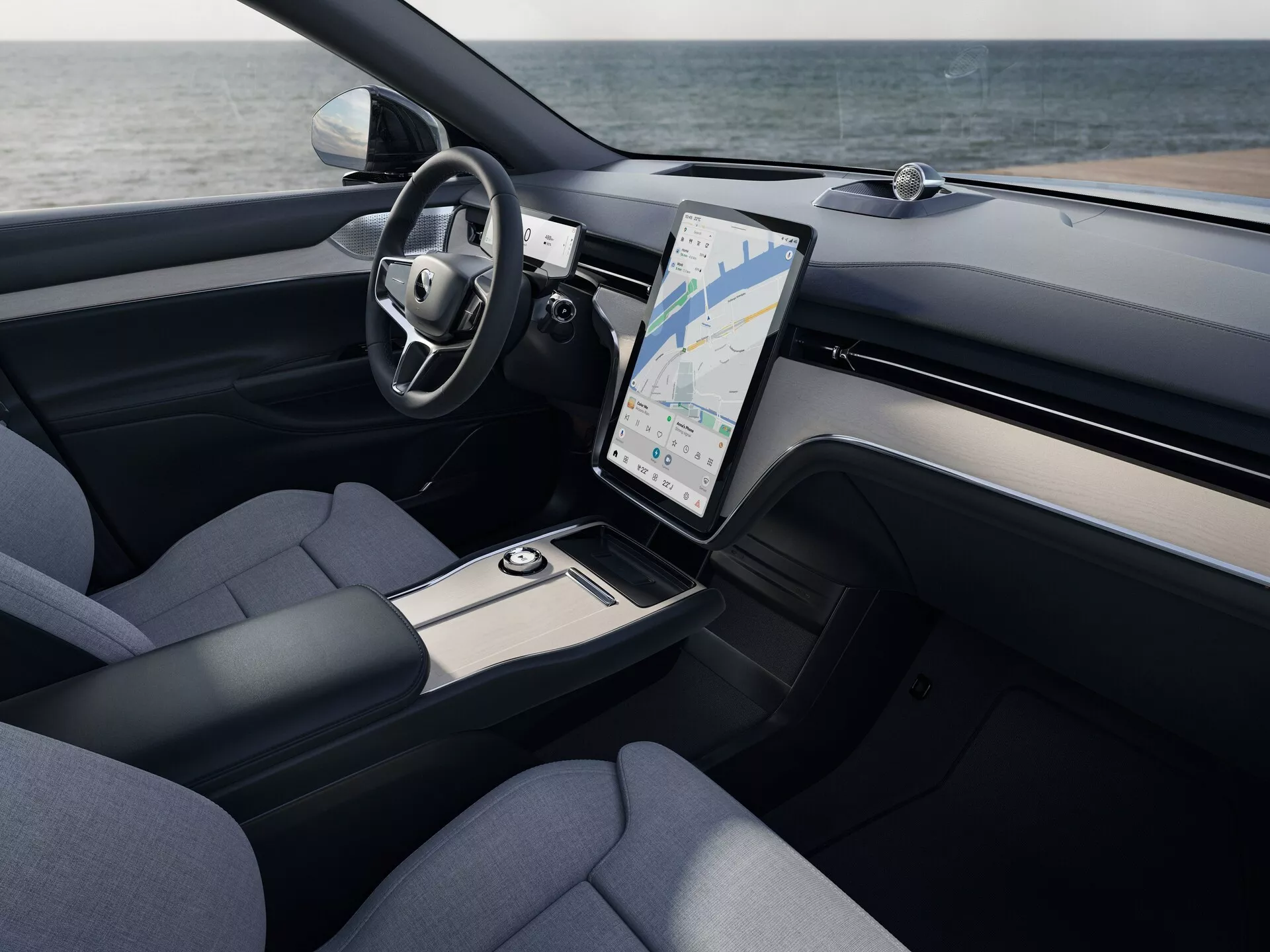 Vue intérieure du Volvo EX90 2023 montrant le coin du conducteur et la console centrale avec une perspective surélevée, présentant un volant multifonction enveloppé de cuir, un grand système d'infodivertissement à écran tactile vertical avec GPS, des bouches d'aération pour le contrôle du climat, des accents de métal brossé, des sièges garnis de tissu et des solutions de rangement élégantes.