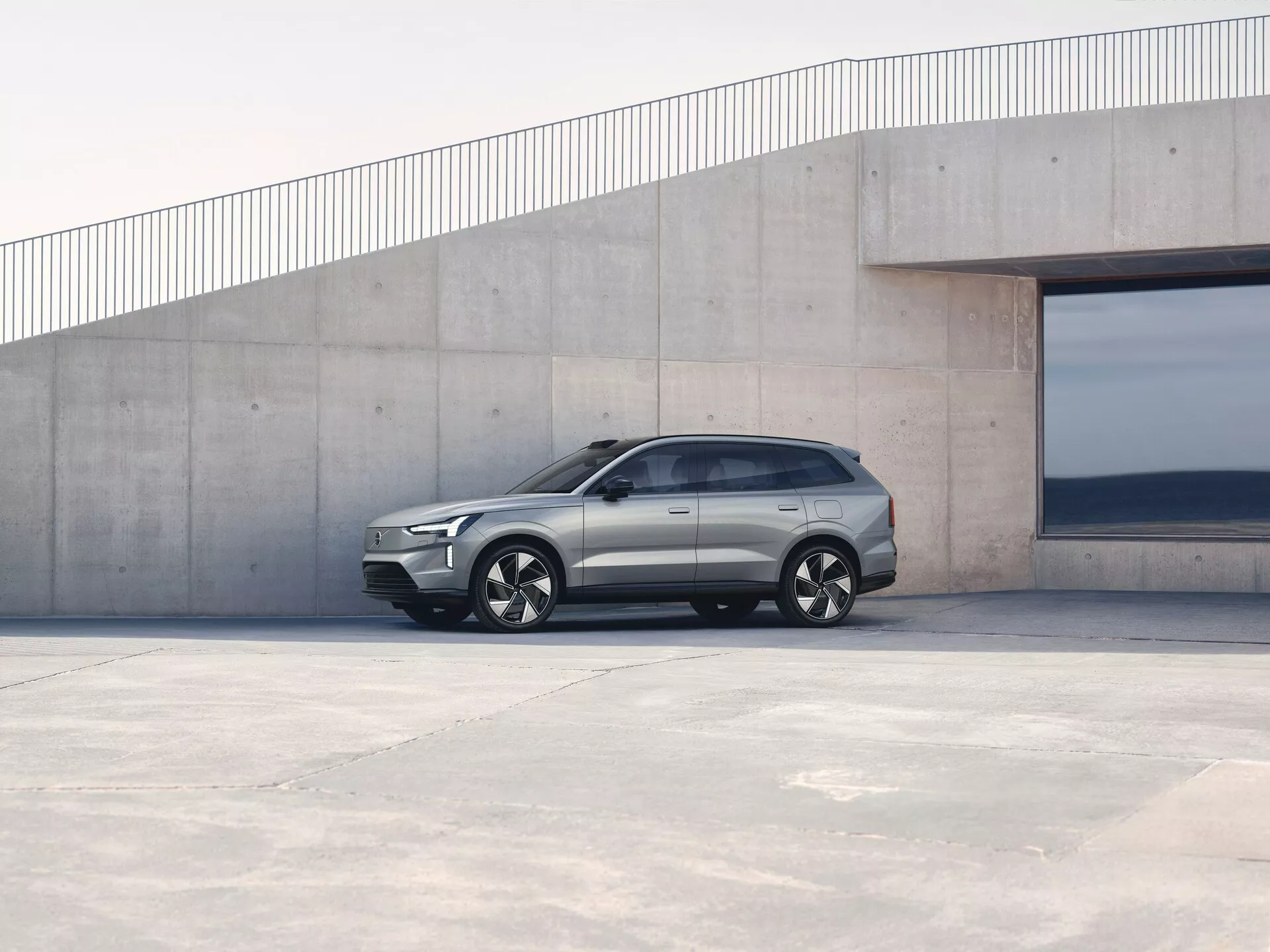 Volvo EX90 de 1ère génération 2023 garée à l'extérieur, vue de profil latéral présentant un design angulaire, des poignées de porte affleurantes et des jantes en alliage contre un fond architectural minimaliste