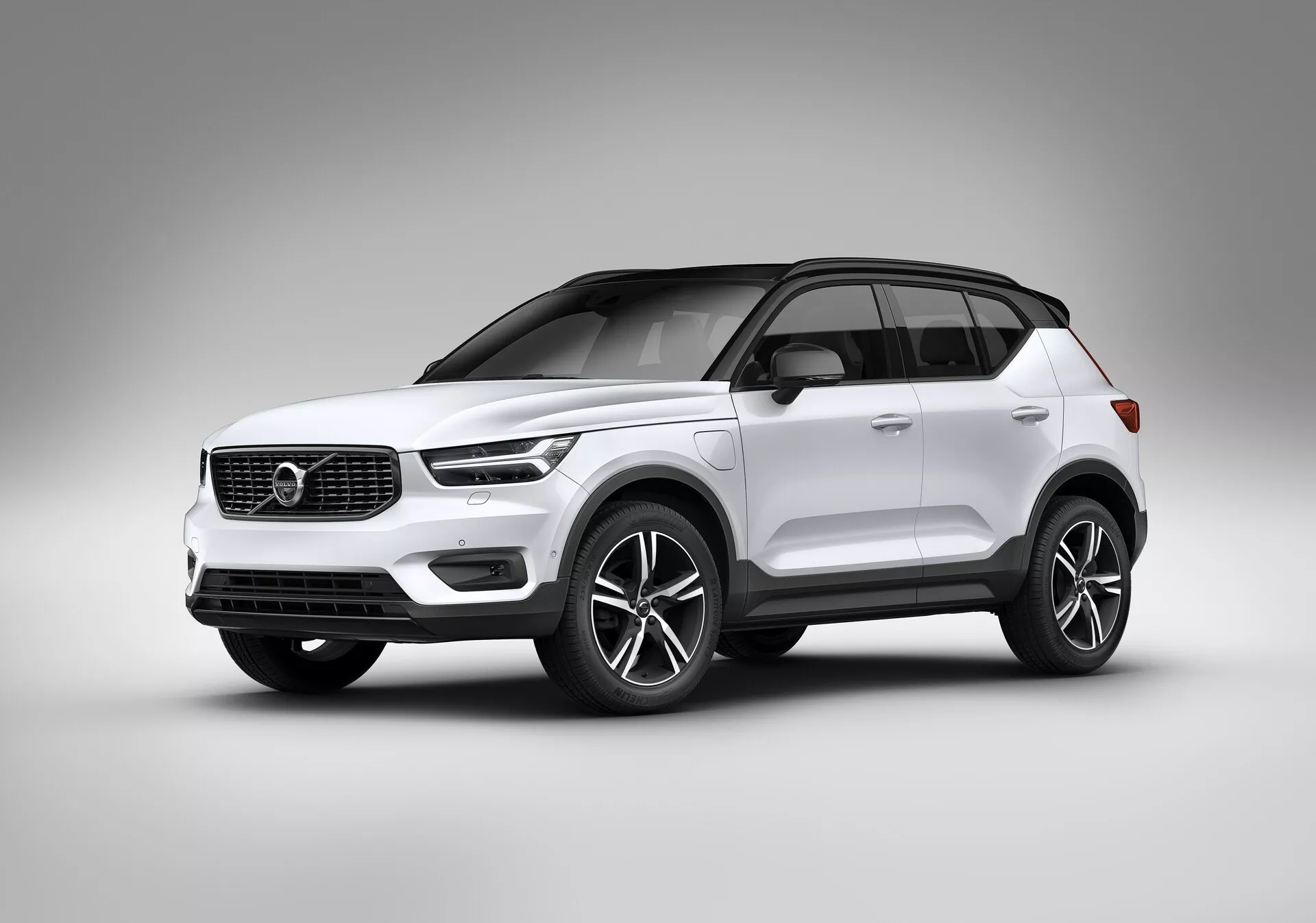 Photographie en studio d'un SUV moderne blanc Volvo XC40 2018 (1ère génération). Prise sous un angle bas, l'image met en valeur la posture et les éléments de design de la voiture, y compris une vue avant trois-quarts montrant les profils avant et latéral. Les phares ont une forme moderne, peut-être avec une technologie LED, tandis que la calandre inclut l'emblème de la marque et des lignes horizontales minimalistes. Le pare-chocs inférieur intègre des feux de brouillard ou des prises d'air, suggérant une technologie avancée. Loules grandes roues en alliage, probablement de 18 pouces, les poignées de portes de la couleur de la carrosserie, les coques de rétroviseurs et le toit panoramique ressortent. La ligne de toit se rétrécit vers l'arrière en style coupé, accentuant le sentiment de luxe avec des garnitures chromées ou métalliques autour des fenêtres. La finition de peinture lisse et brillante et le fond de studio mettent en avant les caractéristiques du véhicule sans distractions, suggérant une attention particulière à la sécurité, le confort, l'infotainment et des systèmes de propulsion avancés.