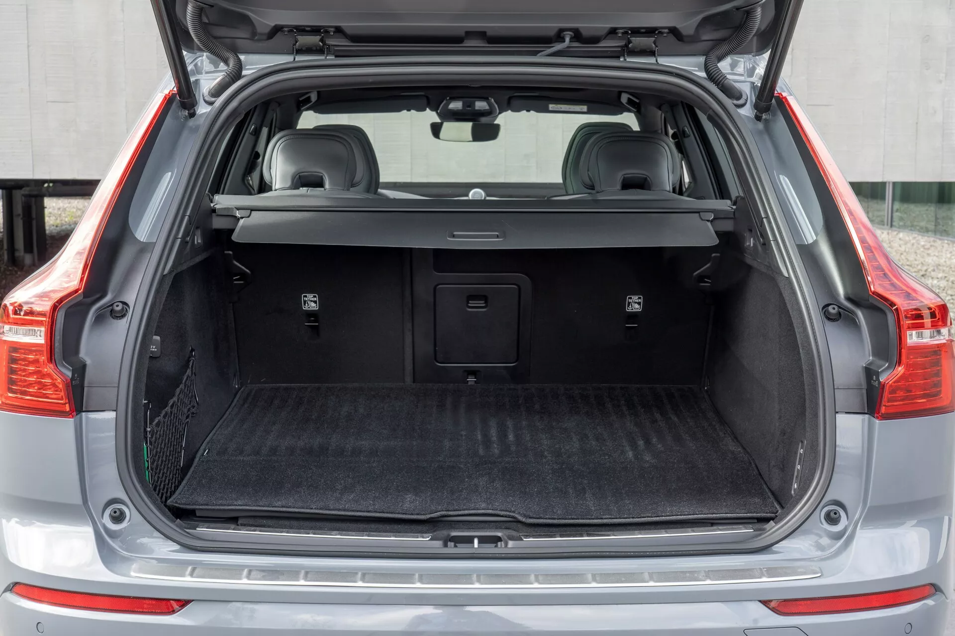 Compartiment de charge arrière d'une Volvo XC60 2021 (2e génération facelift), présentant le coffre ouvert vue arrière droite avec un espace de chargement vide, couvre-bagages rétractable partiellement tiré en arrière, tapis protecteur, garniture plastique avec petits compartiments de rangement, éventuelle prise de courant 12 volts, plaque de seuil en métal et sièges arrière rabattables avec appuie-têtes.
