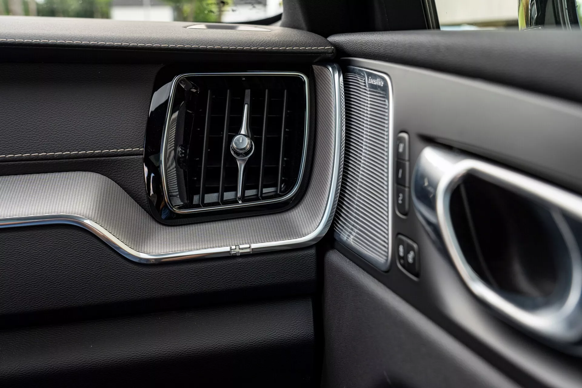 Vue en gros plan du tableau de bord côté passager dans une Volvo XC60 2021 (2ème génération restylée). L'image présente en évidence la bouche de climatisation avec des lamelles horizontales et un bouton rotatif central, encadré par un accent métallique brossé. À droite de la bouche d'aération, il y a une grille de haut-parleur avec des perforations uniformes, à côté de l'accoudoir de la porte et de la poignée partiellement visibles, également ornés de détails métalliques. Le tableau de bord montre des matériaux ressemblant à du cuir dans la partie supérieure et une zone inférieure texturée différemment, séparée par une bande métallique, ce qui met en avant le design premium et la finition de qualité du véhicule.