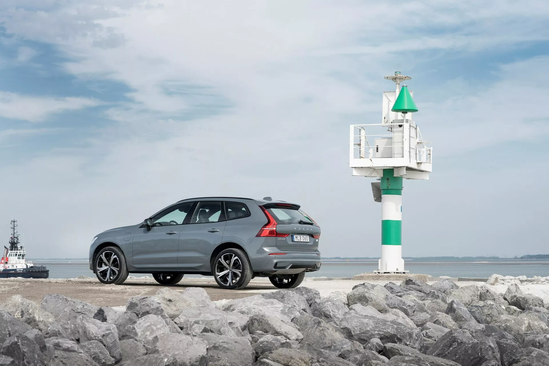 Photographie du modèle Volvo XC60 2021 de deuxième génération restylée, capturée sous un angle arrière trois quarts sur une surface rocheuse, avec un cadre maritime en arrière-plan. La voiture gris foncé à finition métallique présente une silhouette de toit sportive et élégante, des jantes en alliage multi-bâtons, des capteurs de stationnement arrière et une double échappement. À droite de la voiture, un petit phare ou balise est visible, avec un remorqueur ou bateau pilote dans l'eau, ajoutant à l'ambiance côtière.