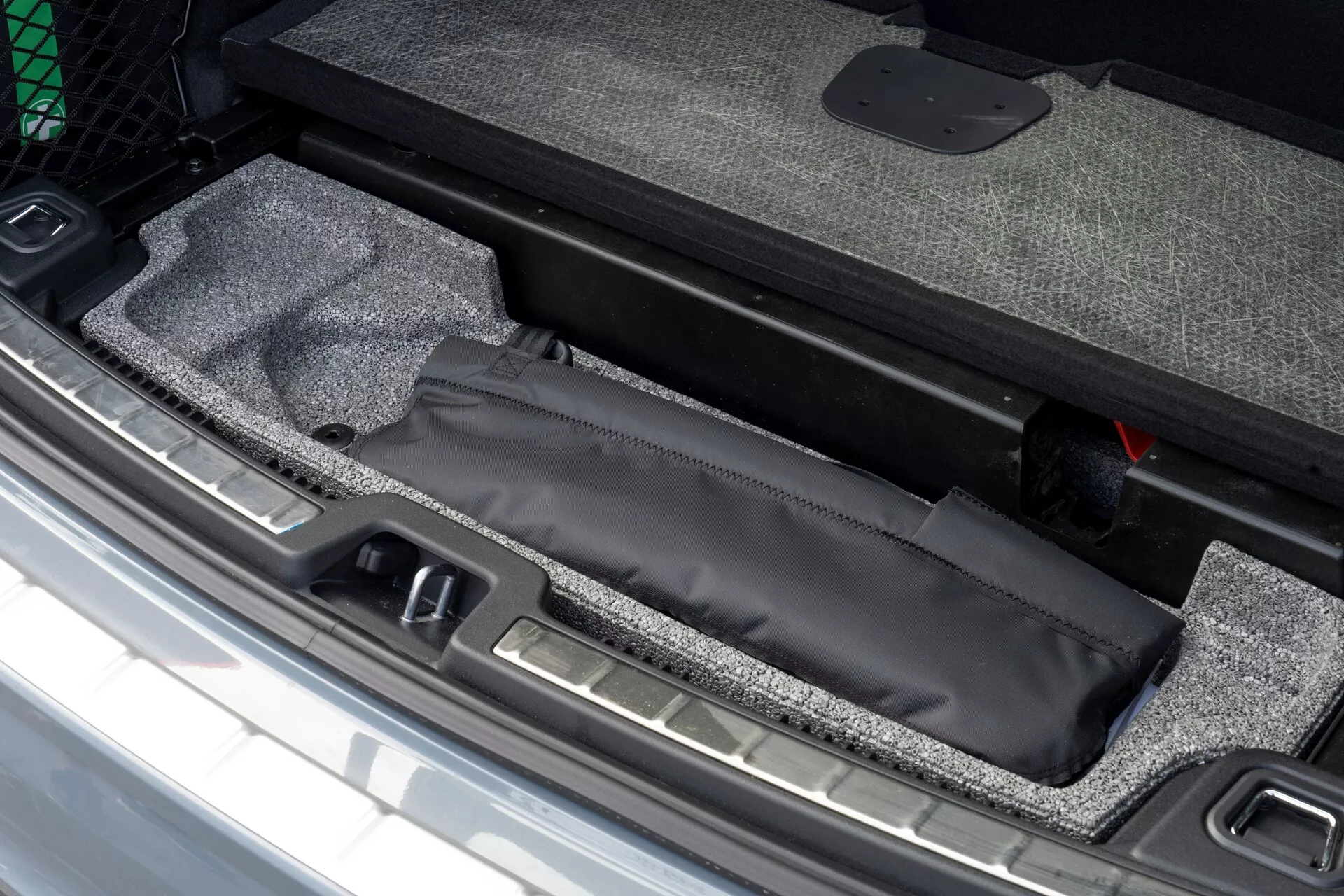 Gros plan de la zone de chargement de la Volvo XC60 2021 montrant un compartiment de rangement caché sous le plancher du coffre avec sac noir, intérieur en moquette et seuil métallique visible, à côté d’un outil d’urgence rouge.