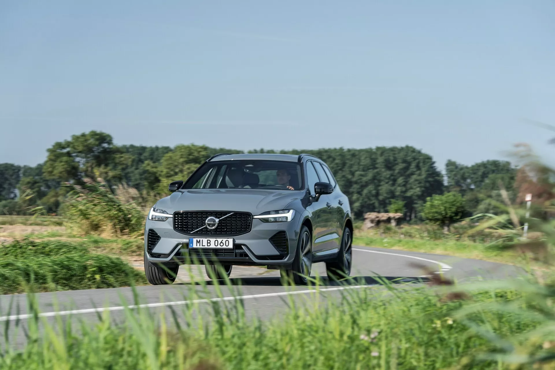 SUV moderne, Volvo XC60 2021 (restylage de 2e génération), capturé sous un angle trois-quarts avant bas, mettant en valeur la posture dynamique et les caractéristiques de design. Design de la calandre proéminente avec grand emblème, phares LED élégants et pare-chocs sculpté avec prises d'air intégrées. Apparence aérodynamique et haut de gamme avec des lignes profilées, une grande garde au sol, de grandes jantes en alliage à plusieurs branches, des pneus à profil bas, des rétroviseurs latéraux avec indicateurs de virage intégrés, une ligne de toit en pente et des vitres teintées. Suggère des fonctionnalités technologiques et de sécurité avancées, conduisant sur une route rurale avec une végétation floue suggérant le mouvement.