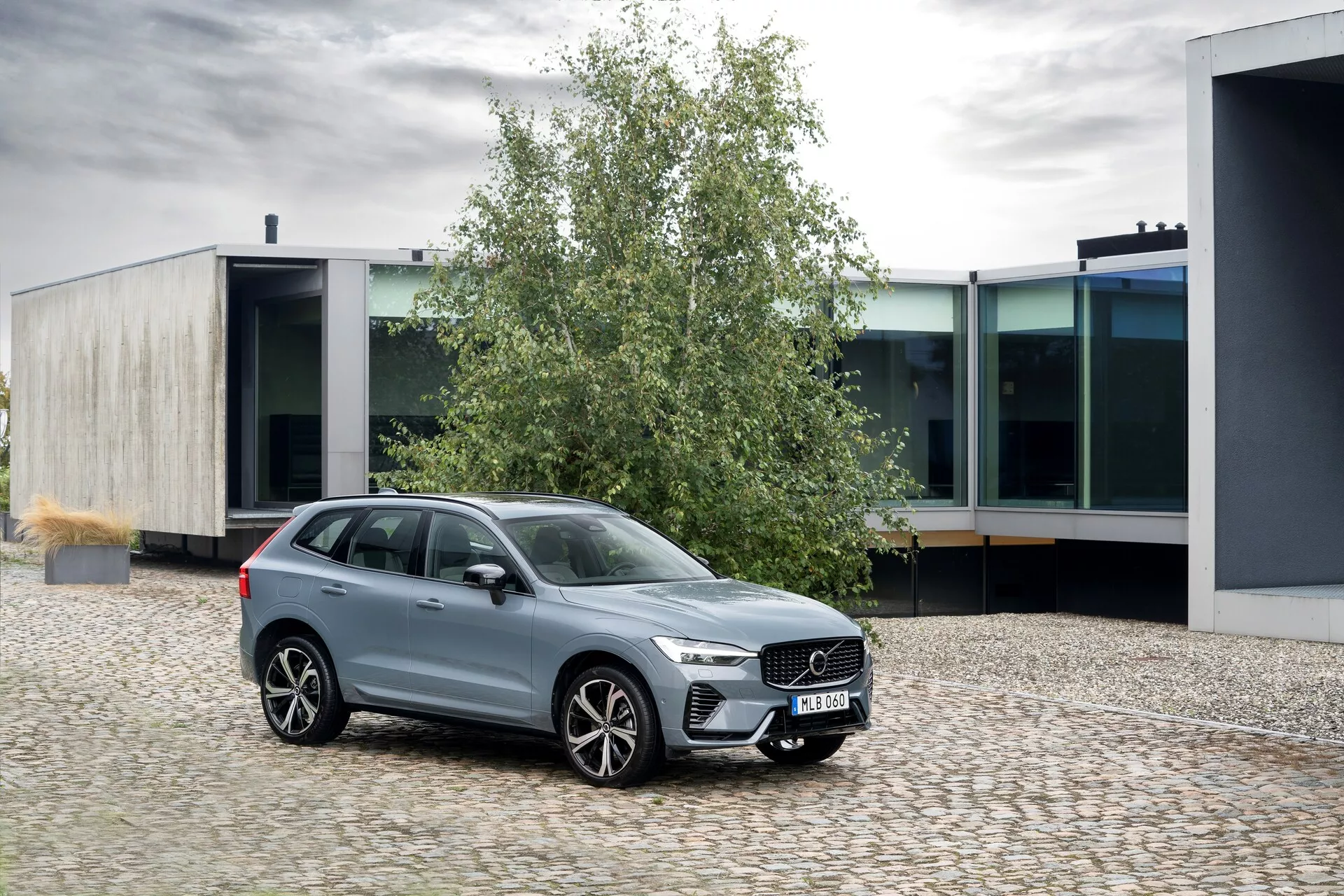 Image d'un Volvo XC60 2021 (2e génération-restylage) garé sur une surface en pavés, positionné à un léger angle vers la gauche. La perspective trois-quarts avant met en lumière sa grille imposante avec des barres verticales, ses phares LED élégants avec feux de jour, et ses grandes jantes en alliage à plusieurs rayons. Les fenêtres sont entourées de chrome, rehaussant sa finition métallique polie. Le SUV présente une garde au sol élevée, des barres de toit et des rétroviseurs aux couleurs de la carrosserie avec des clignotants intégrés. La scène se situe dans un environnement urbain avec une architecture moderne, soulignant son design élégant et ses caractéristiques de luxe.
