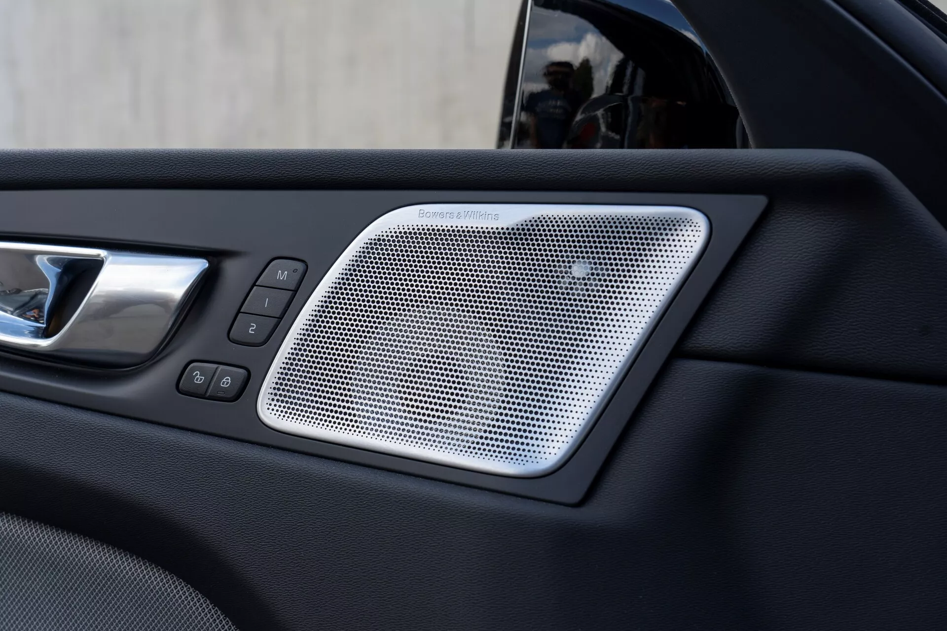 Image en gros plan de la section intérieure de la porte d'une Volvo XC60 2021 (2e génération, facelift) montrant une grille de haut-parleur Harman/Kardon au design en maille, une poignée de porte chromée et des commandes de verrouillage et de fenêtres électriques, mettant en valeur le luxe et la modernité des caractéristiques du véhicule.