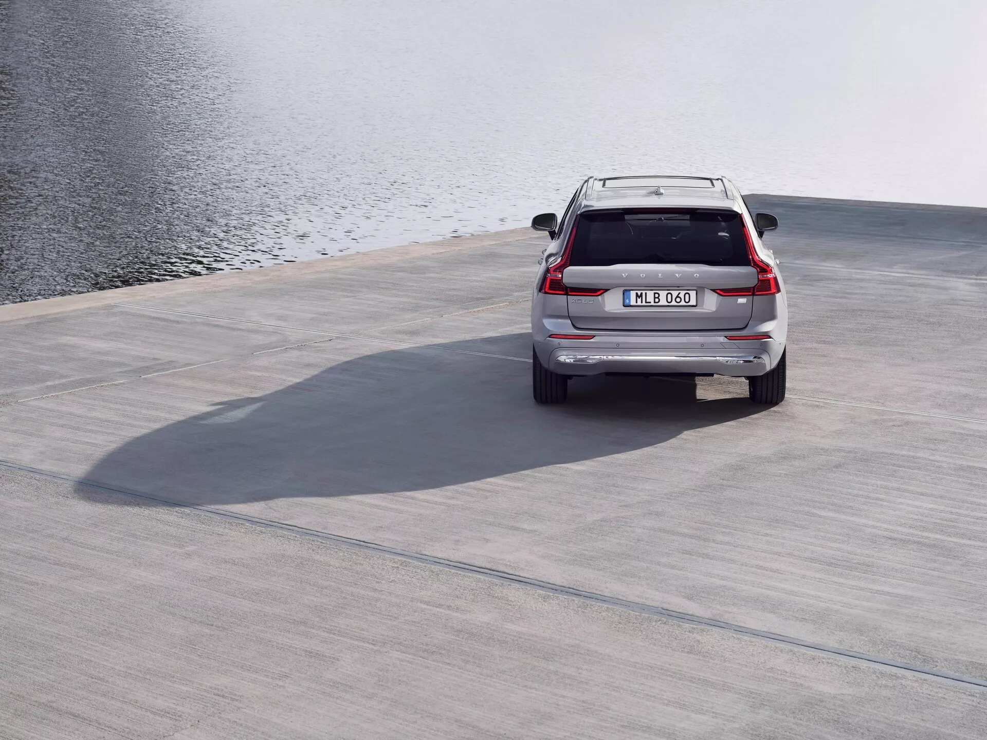 Vue arrière d'un SUV Volvo XC60 2021 (deuxième génération, facelift) argenté, garé sur une surface en béton près d'un plan d'eau, montrant le becquet de toit, les feux arrière à LED, le hayon, le pare-chocs arrière et les tuyaux d'échappement vus d'un angle élevé avec des ombres projetées, dans des conditions calmes et de vent léger.