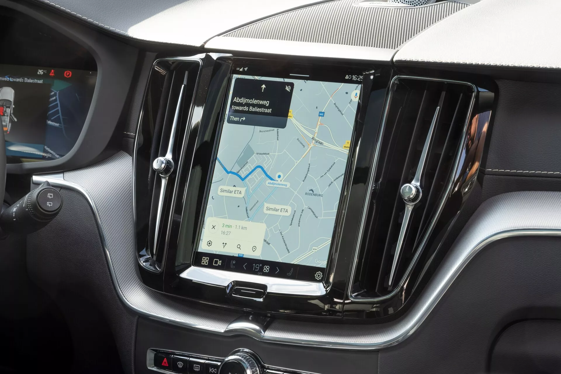 Gros plan de la console centrale d'une Volvo XC60 2021 (2e génération, restylée), centrée sur le système d'infodivertissement. La photo, prise depuis la perspective du passager avant, présente un grand écran tactile vertical affichant une carte de navigation. L'écran est encadré par une garniture métallique et entouré d'une texture semblable à la fibre de carbone, mettant en valeur des éléments de design intérieur modernes et sportifs. Les boutons physiques sont visibles sur le côté de l'écran et en dessous pour le contrôle de la climatisation, indiquant une combinaison de fonctions tactiles et physiques. La vue partielle du volant sur le bord gauche confirme une configuration de conduite à gauche.
