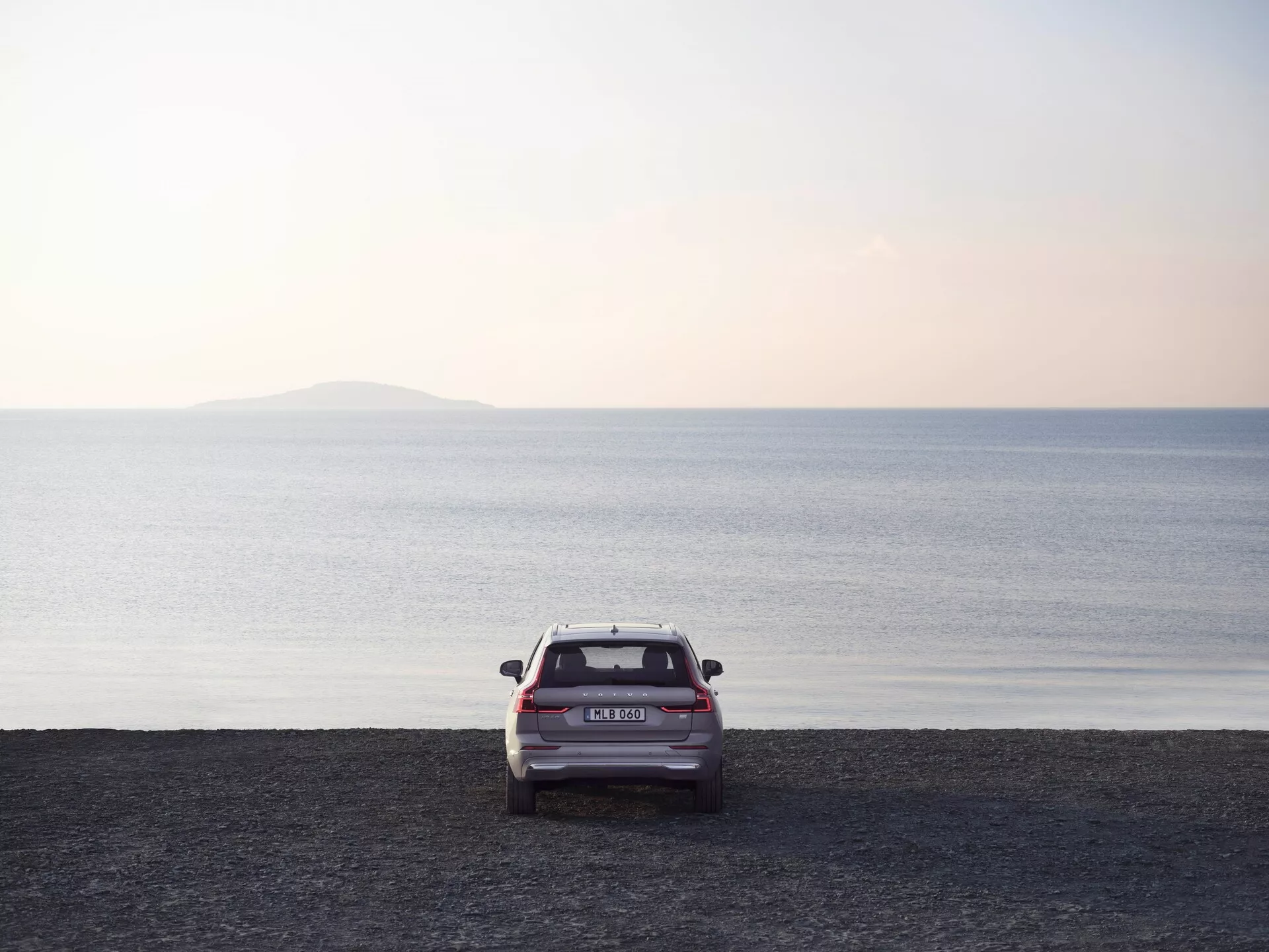 Vue arrière d'un Volvo XC60 2021 de deuxième génération facelift, garé sur un rivage couvert de galets avec une mer calme et un horizon dégagé, capturée au lever ou au coucher du soleil avec une lumière douce. Les feux arrière, le pare-brise arrière, le coffre, le pare-chocs arrière et les barres de toit du SUV sont visibles, soulignant son design moderne et sa praticité. La photo met en avant le véhicule placé dans un vaste cadre côtier serein avec un léger reflet sur les galets mouillés, indiquant une récente activité de marée.