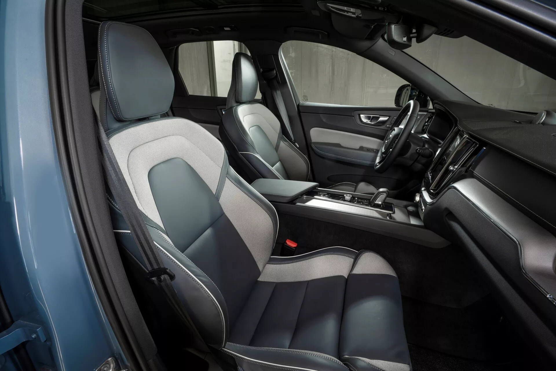 Intérieur du Volvo XC60 2021 (2e génération-restylage) photographié depuis un angle oblique surélevé du côté conducteur, mettant en valeur le design et les caractéristiques de l'habitacle avant, y compris des sièges en cuir et tissu gris et noir, une console centrale avec levier de vitesse et divers contrôles, un tableau de bord avec écran tactile d'infodivertissement, continuité dans le design des panneaux de porte et grilles de haut-parleurs visibles.