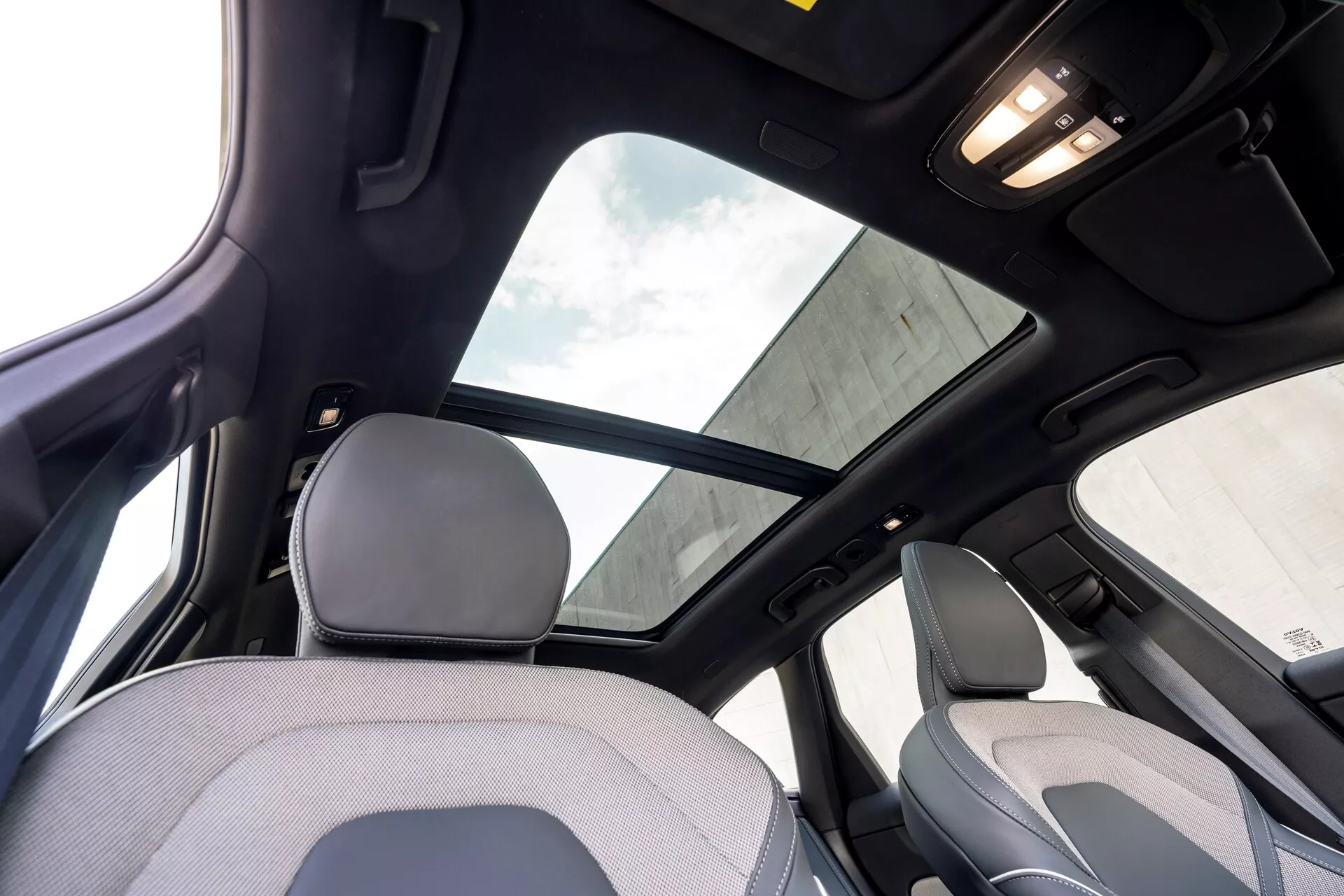 Intérieur du Volvo XC60 2021, de la 2e génération restylée, vue depuis le siège arrière, se concentrant sur la partie supérieure de l'habitacle. Les caractéristiques notables incluent un grand toit ouvrant panoramique avec pare-soleil, ciel de toit noir indiquant un niveau de finition élevé ou un pack de luxe, et éclairage de la cabine au plafond. La prise de vue capture une partie de l'appuie-tête et du dossier supérieur du siège passager avant, ainsi que le montant B et la poignée au-dessus de la porte arrière.