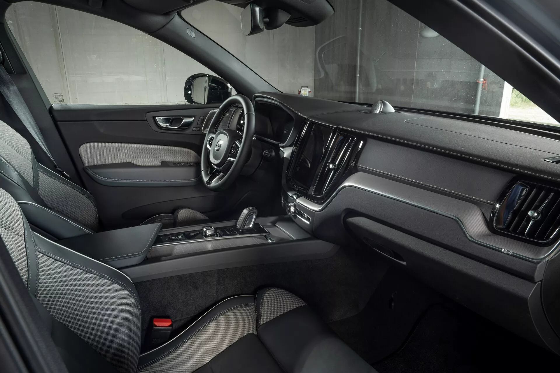 Vue intérieure de la Volvo XC60 2021 (facelift de 2ème génération) depuis le côté passager avant, mettant en valeur la porte du conducteur avec des systèmes de commande électronique, volant à trois branches avec boutons multifonctions, grand écran tactile central pour l'infodivertissement et design luxueux avec cuir foncé et accents métalliques.