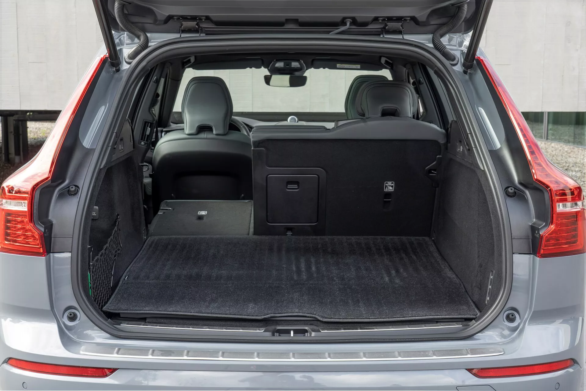 L'image présente l'aire de chargement arrière du Volvo XC60 2021 (2ème génération avec lifting), vue de face avec le coffre ouvert. Le plan à distance moyenne capture toute la largeur de l'espace de chargement, avec un plancher de chargement plat recouvert de matériau type moquette noire. Le côté gauche comprend un petit compartiment de rangement avec un filet. Le siège arrière droit est rabattu, révélant la capacité de pliage fractionnée, tandis que le siège gauche reste droit avec un appui-tête. Une couverture de charge rétractable se trouve au-dessus des sièges arrière, illustrant la polyvalence de stockage du véhicule.
