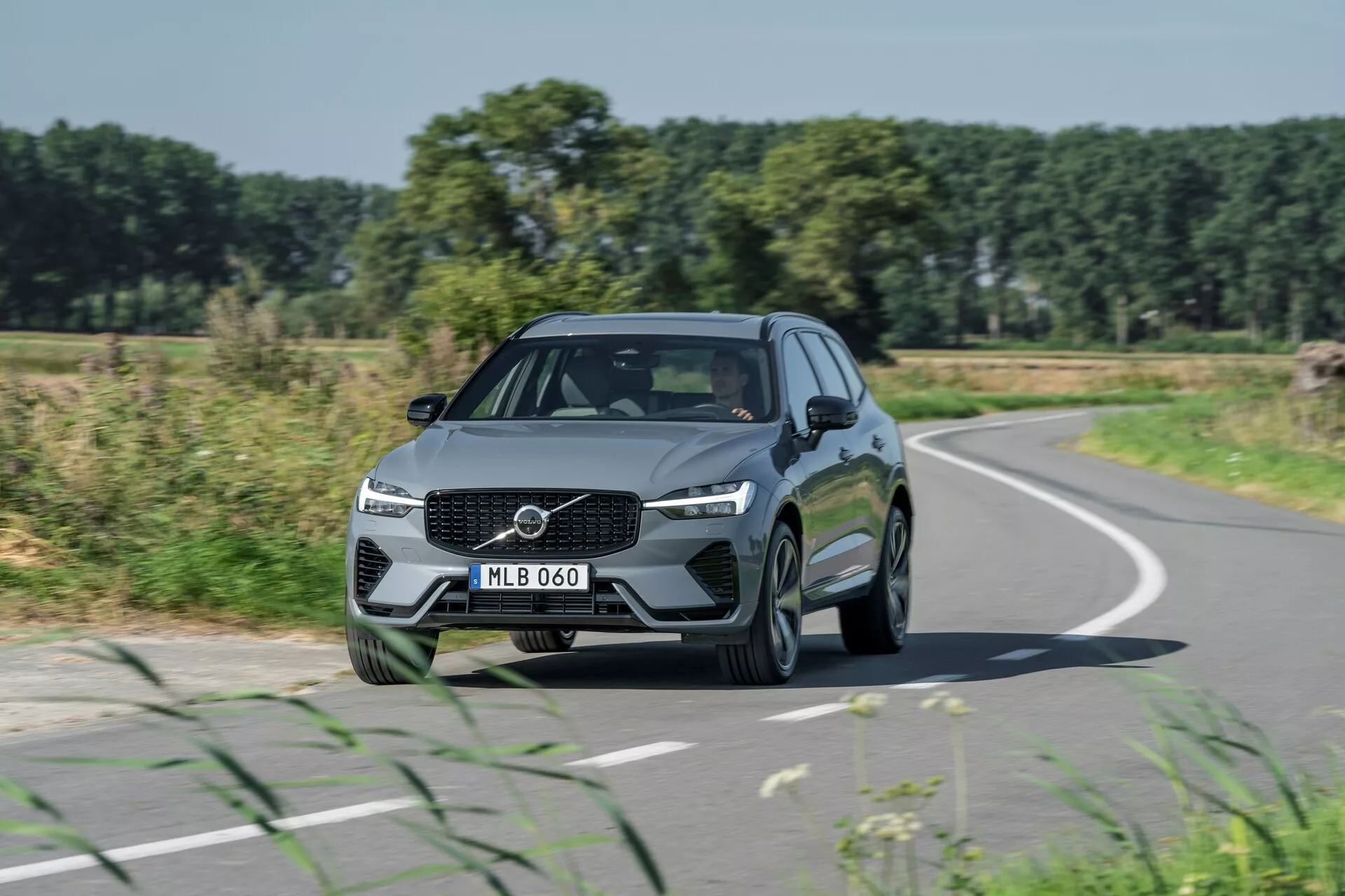 L'image capture une Volvo XC60 2021 (facelift de 2e génération) en mouvement sous un angle de trois quarts avant, mettant en avant des éléments de design tels qu'une calandre proéminente avec logo centré, des phares LED angulaires et des lignes élégantes sur le capot. La voiture, circulant sur une route légèrement courbée, présente une teinte métallique et des jantes en alliage à multiples rayons, flanquées de lignes contournées et de rétroviseurs latéraux intégrés avec clignotants. Avec en arrière-plan des champs, des arbres et un ciel bleu clair, cette représentation met en avant le design automobile, les voyages routiers et les scénarios de conduite réels.