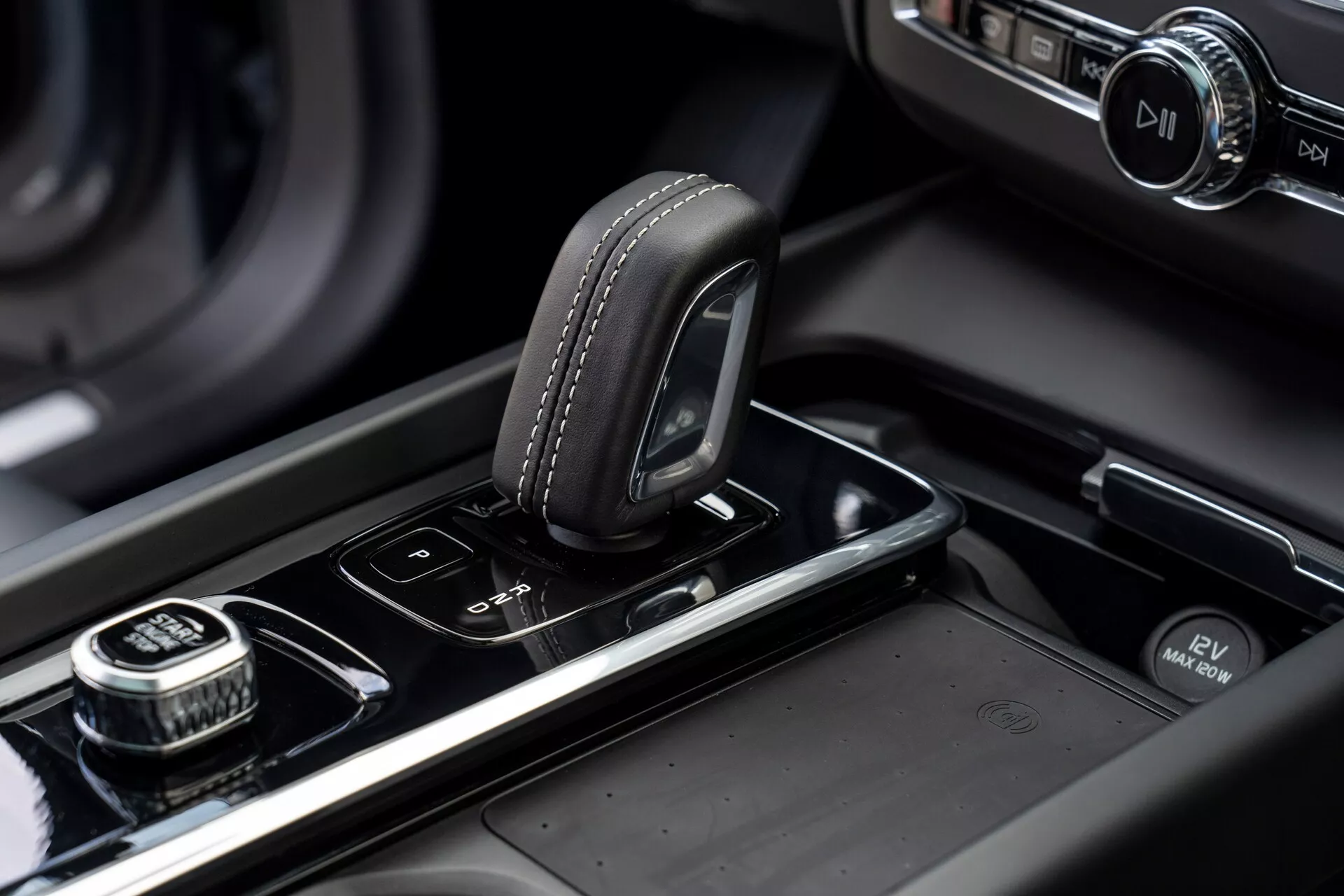 Gros plan de la console centrale d'une Volvo XC60 2021 (2e génération, facelift). L'image met en avant le sélecteur de vitesses avec une partie supérieure en cuir et des détails de couture, situé sur une surface noire brillante. À droite se trouve une molette à bord cranté, probablement pour la navigation dans le système d'infodivertissement, avec un bouton de sélection par pression. À côté du sélecteur de vitesses se trouvent des boutons pour les capteurs de stationnement et le système d'arrêt/démarrage automatique. La console centrale comporte des garnitures métalliques et un écran d'infodivertissement illuminé en arrière-plan flou, soulignant le design moderne et haut de gamme du véhicule ainsi que son artisanat.