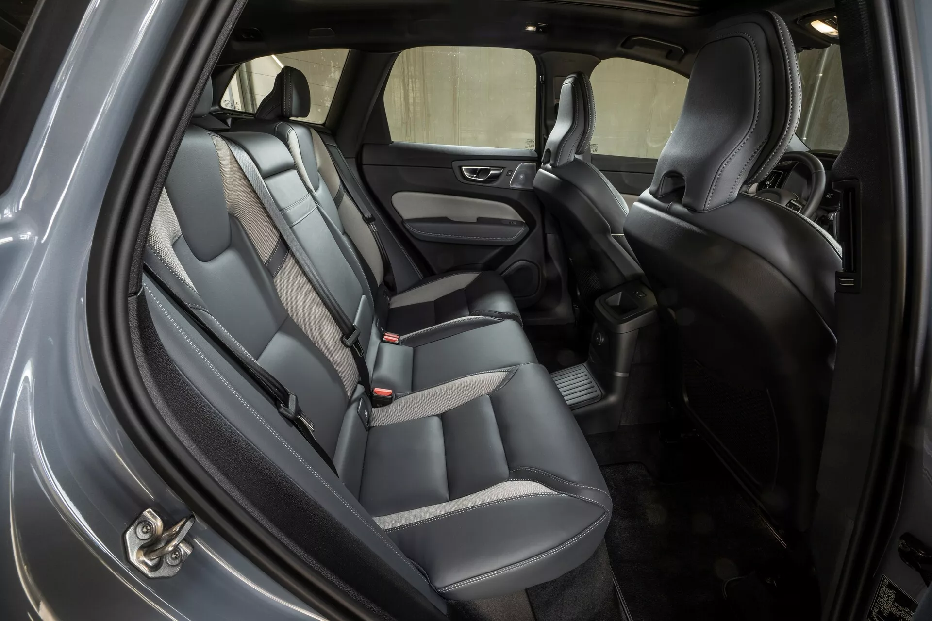 Vue intérieure de la Volvo XC60 2021, montrant la zone des sièges arrière sous un angle oblique avec la porte arrière du côté trottoir ouverte. La voiture est équipée de sièges en cuir ou similicuir bicolores, d'appuie-têtes, de boucles de ceinture de sécurité pour trois passagers, et de panneaux de porte garnis avec un haut-parleur intégré et des commandes de vitres électriques. Le sol est recouvert de moquette avec un tapis de sol et la console centrale visible entre les sièges avant comprend un espace de rangement. L'habitacle bien éclairé suggère un éclairage ambiant et supérieur, offrant une bonne visibilité à travers les fenêtres latérales et arrière.