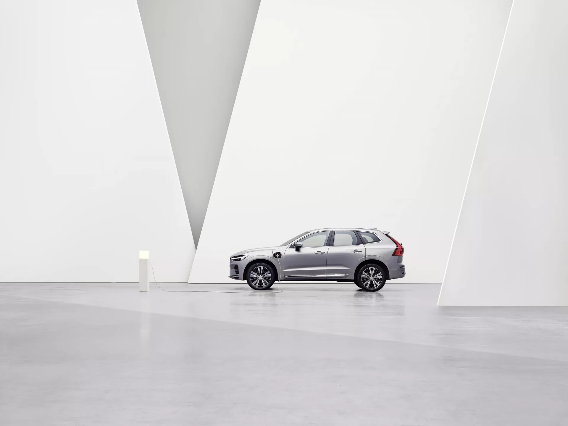 L'image présente un SUV Volvo XC60 2021 de deuxième génération avec lifting, dans une galerie d'art spacieuse et minimaliste avec des murs blancs et un sol brillant. Le véhicule, vu de profil et orienté vers la gauche, met en avant les jantes en alliage, les lignes de caractère sur les portes latérales, les rétroviseurs, les vitres teintées et les barres de toit distinctives. D'un angle légèrement bas, on peut voir la calandre avant, les phares LED, les clignotants intégrés et les capteurs de stationnement du pare-chocs avant. L'environnement propre et la paroi en forme de V mettent en valeur le design de la voiture et sa position premium sur le marché sous un éclairage doux et diffus.