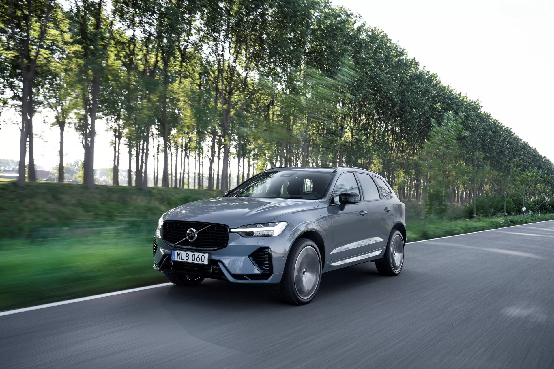 L'image présente une Volvo XC60 2021 (facelift de 2ème génération) en mouvement, capturée sous un angle de vue de trois quarts avant, montrant le côté conducteur de la voiture. Ce SUV moderne et luxueux a un design élégant et un emblème sur la grille avant. La photo, prise sous un léger angle descendant, inclut le capot, le pare-chocs, la grille, les phares et les roues. Le véhicule de couleur métallique foncée, peut-être gris ou noir, avec une finition brillante, dispose de jantes en alliage, de phares LED et d'une carrosserie aérodynamique. L'arrière-plan flou d'un paysage urbain avec des arbres suggère le mouvement et met en valeur les capacités de performance de la voiture.