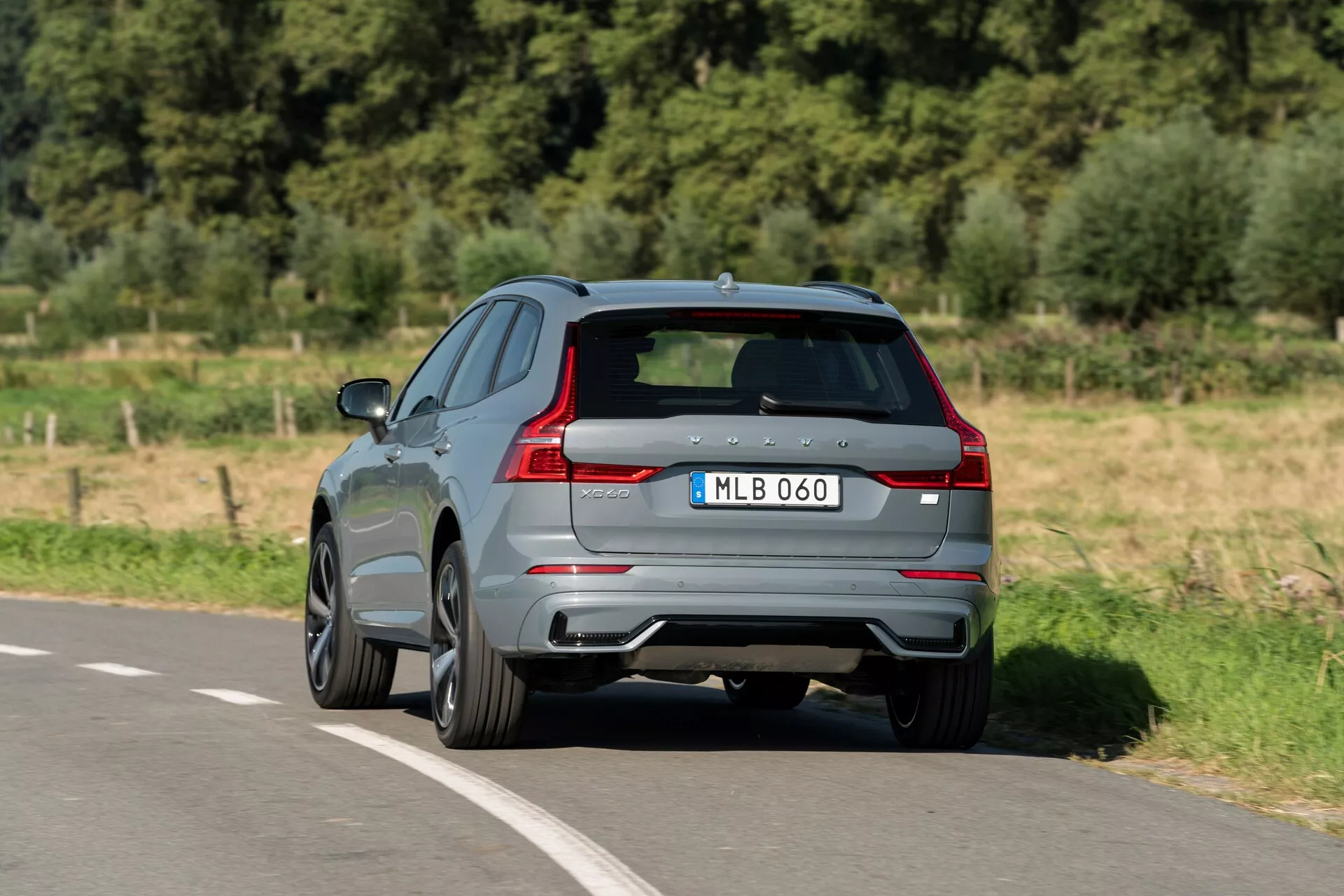 Vue arrière trois quarts d'un Volvo XC60 2021 circulant sur une route pavée. Capturée sous un angle élevé, ce SUV moderne met en valeur sa peinture grise brillante, ses feux arrière LED distinctifs, ses doubles sorties d'échappement et son diffuseur arrière. Le badge du fabricant et la désignation du modèle sont visibles sur le hayon sculpté, complétés par des vitres teintées et des jantes en alliage au design multi-rayons, suggérant une finition plus sportive. L'image offre une perspective claire du design aérodynamique et des caractéristiques haut de gamme propres à un véhicule moderne.